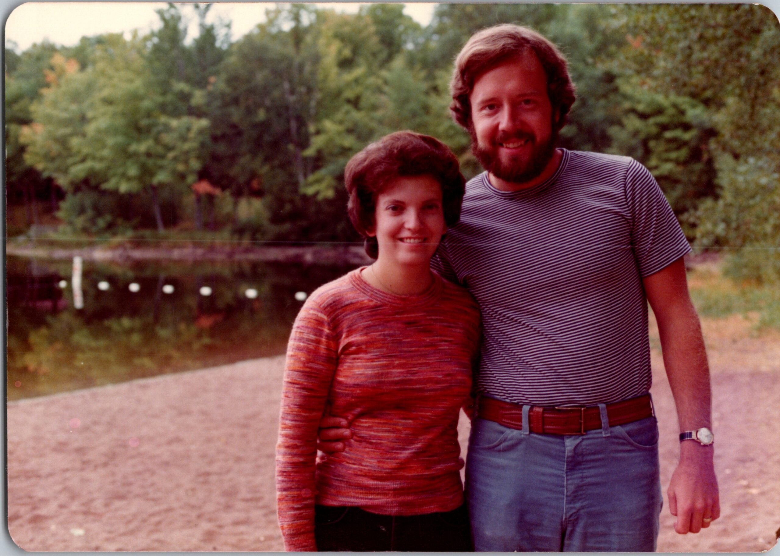 Two people posing outdoors with trees in the background—a man with a beard in a striped shirt stands beside a woman in a red striped sweater, both smiling and facing the camera.