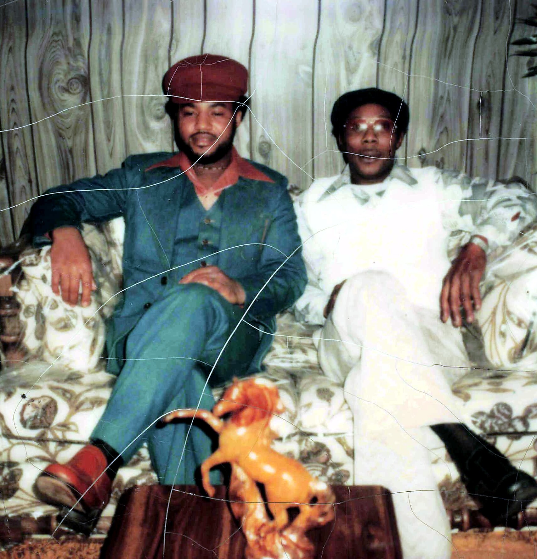 Two men sit on a floral patterned sofa in a retro living room; one wears a teal suit and red hat, the other a white suit with glasses, with an orange toy in front.