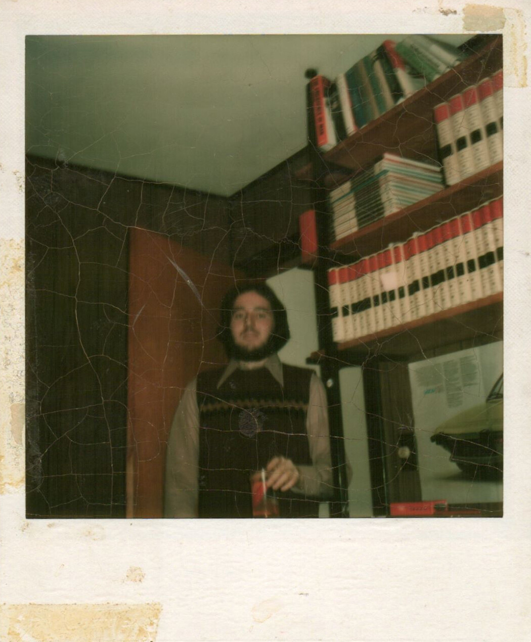 Bearded man in a sweater vest and glasses stands in a room with shelves of white binders behind him, holding a red can.