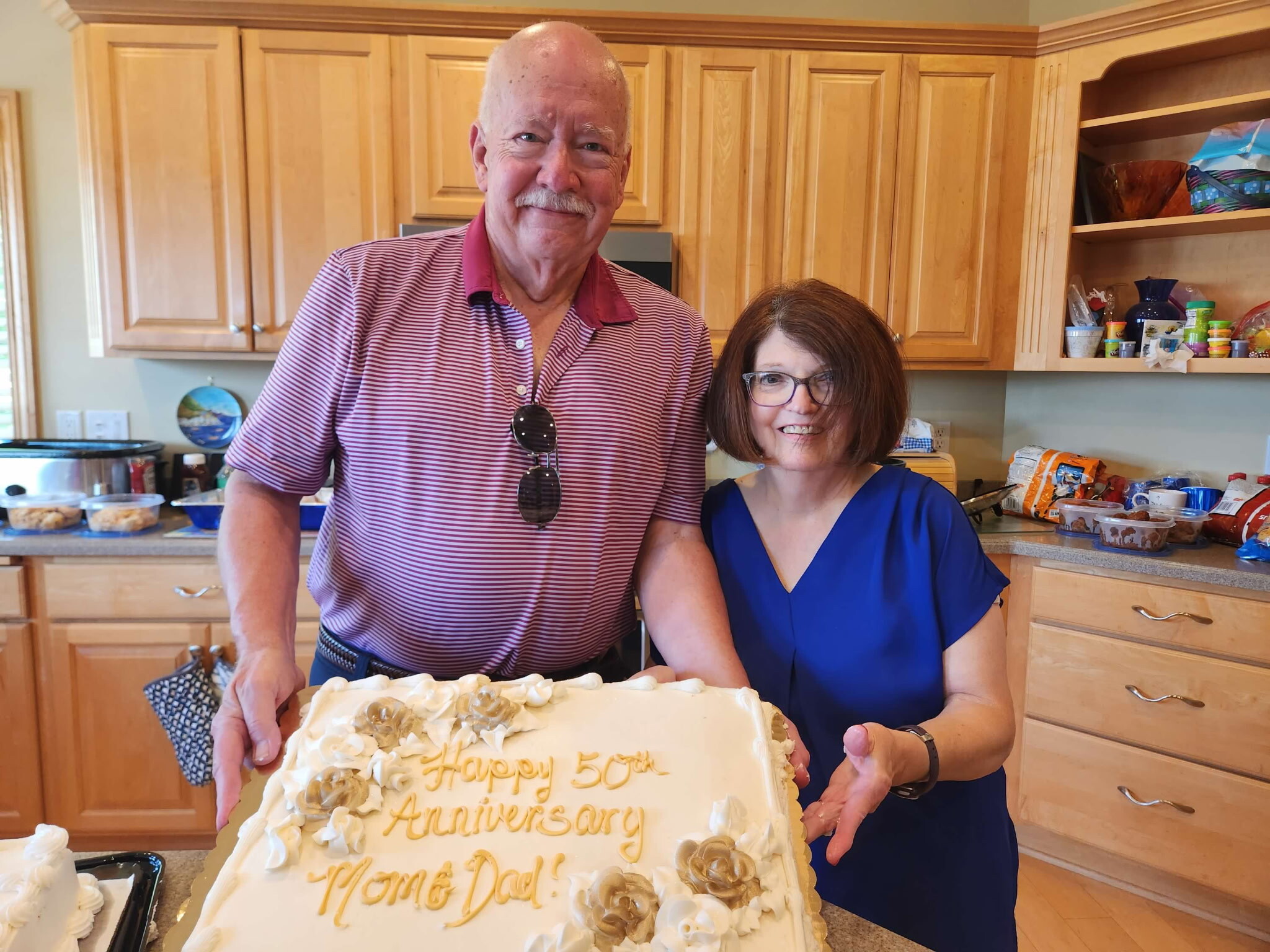 Elderly man and woman smile while presenting a large decorated cake in a bright kitchen.