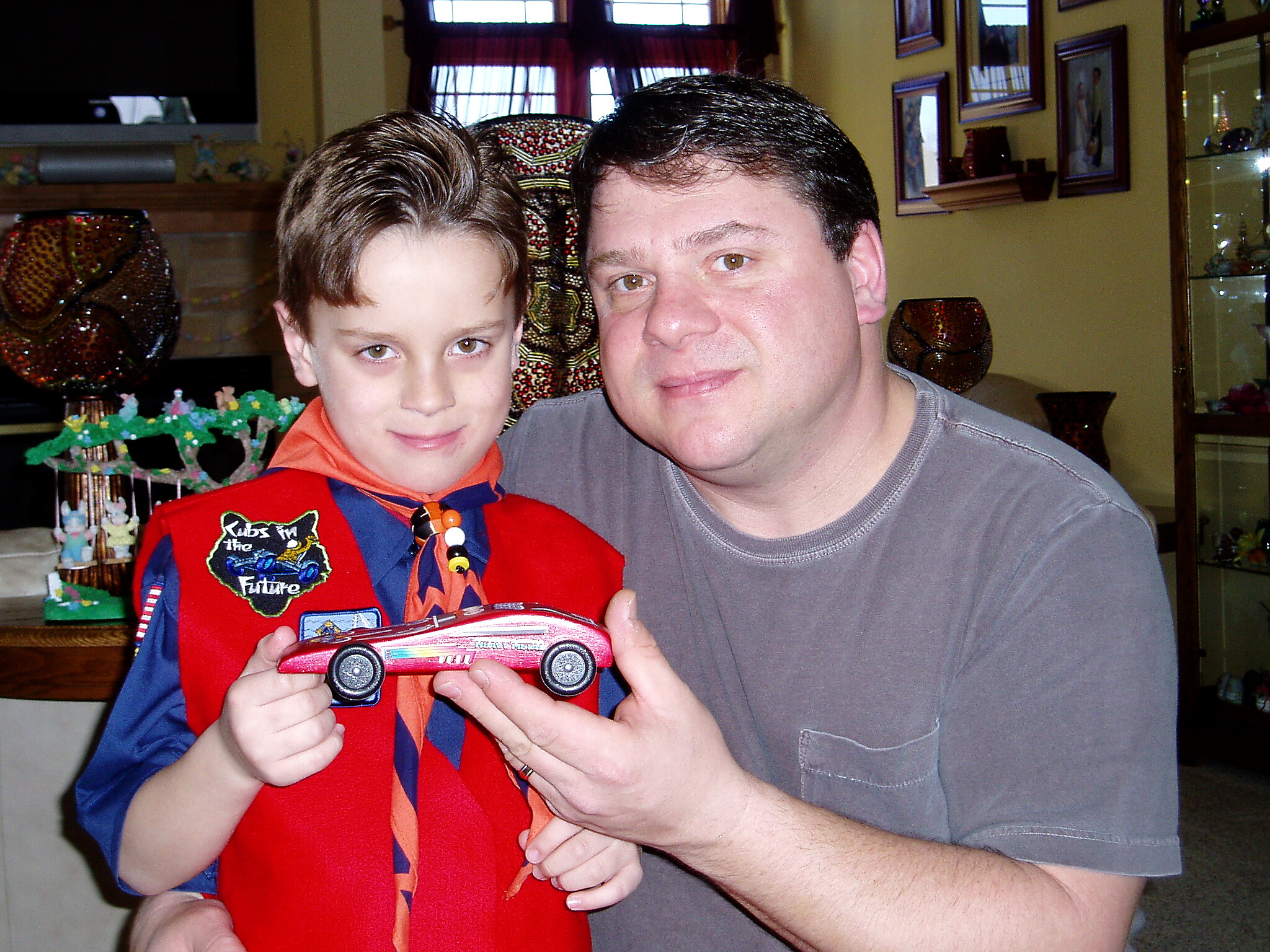 Adult and child pose with a red toy car in a home setting, colorful decorations in the background, square windows visible behind them.