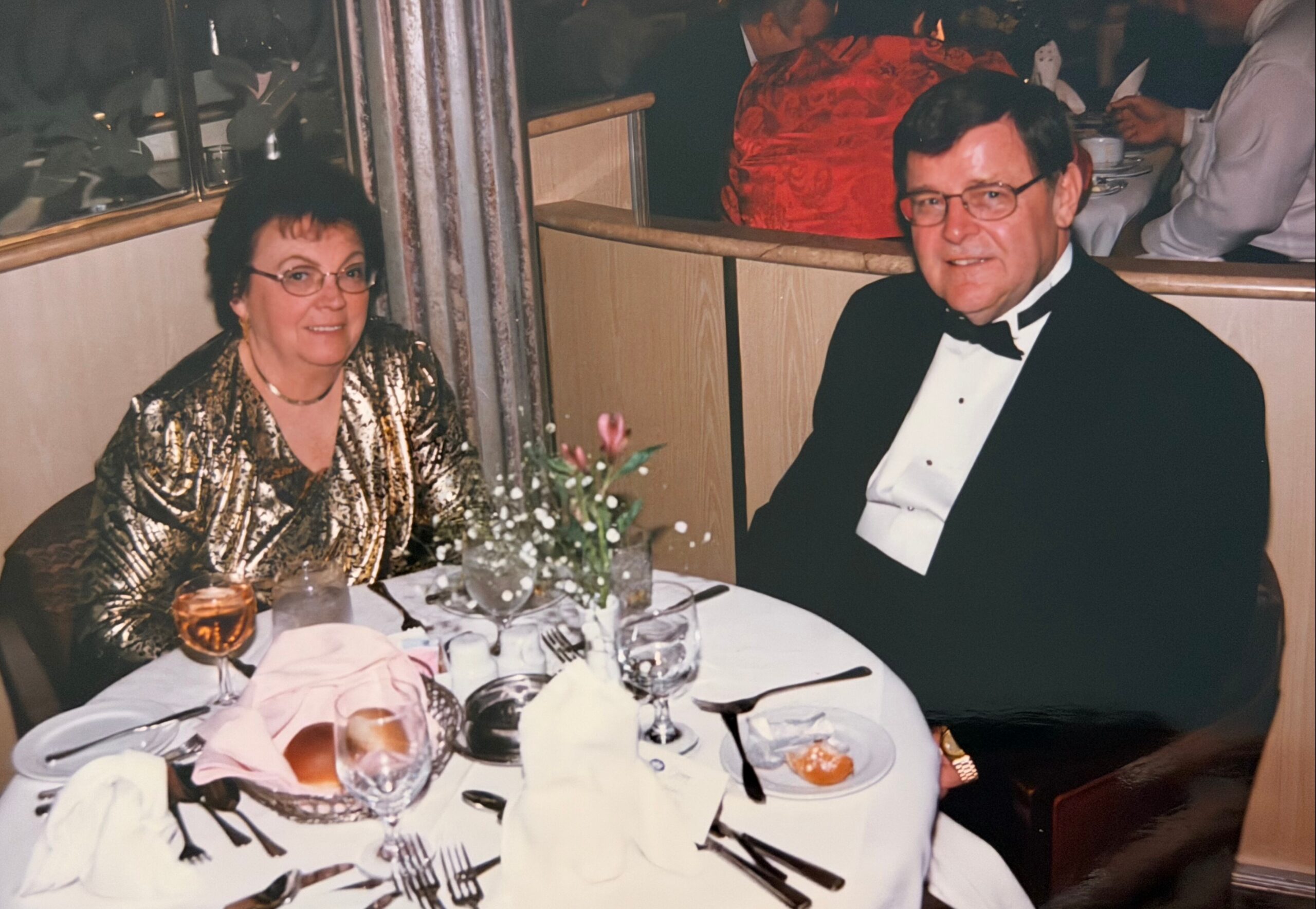 Elegant couple at a formal dinner, woman in a gold metallic dress and man in a black tuxedo, seated at a table with wine glasses and a floral centerpiece.