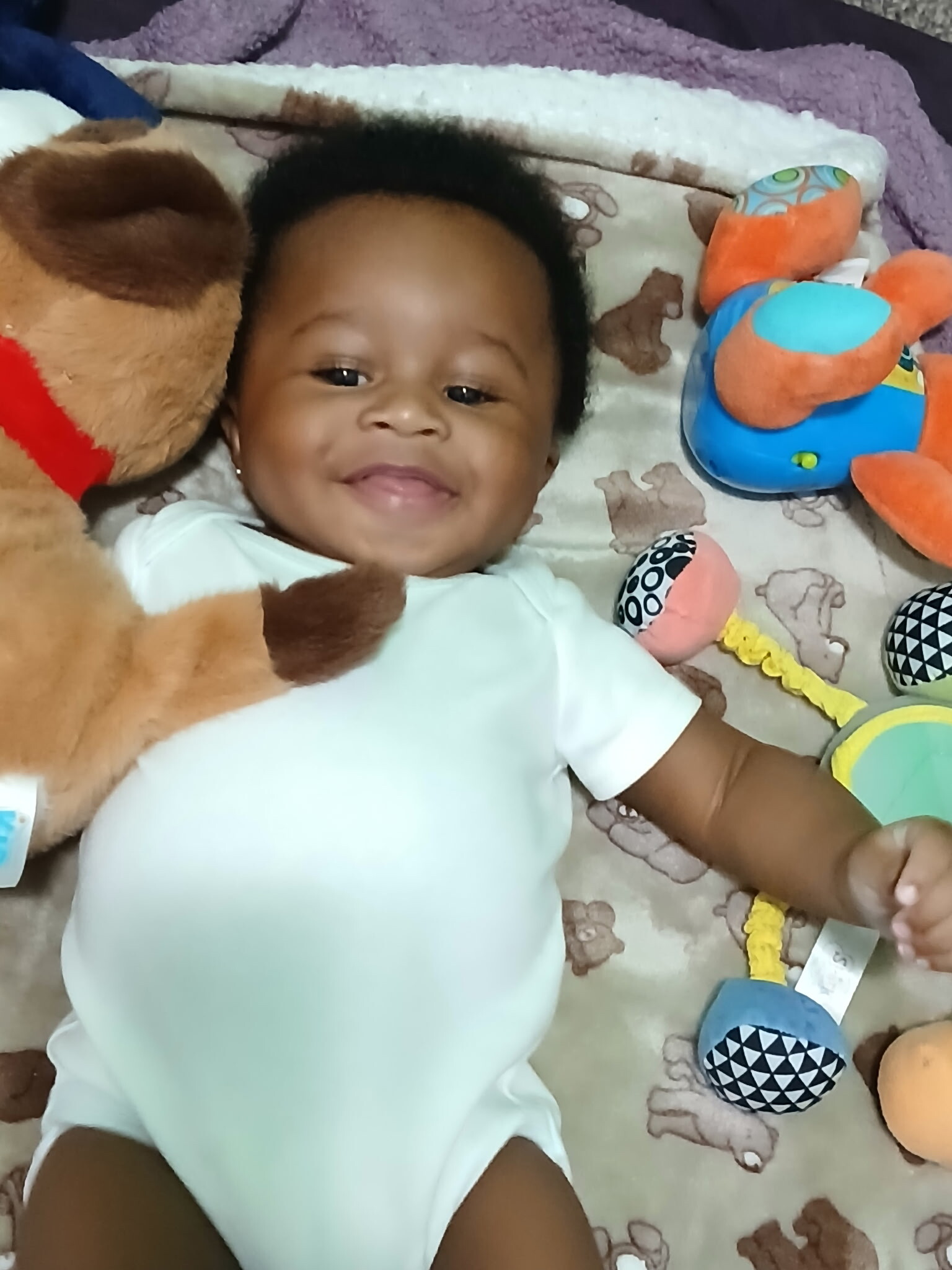 Smiling baby lying on a blanket surrounded by stuffed animals and soft toys at playtime (teddy bear to left).