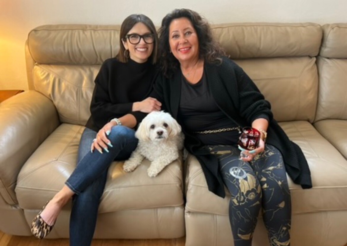 Two women sit closely on a beige sofa, with a small white dog resting on the lap between them, all smiling at the camera.