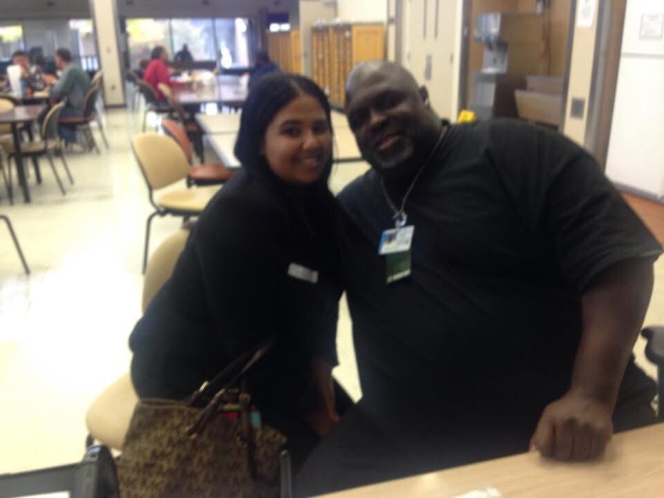 Two people sit together in a school cafeteria, smiling at the camera with tables and other diners in the background.