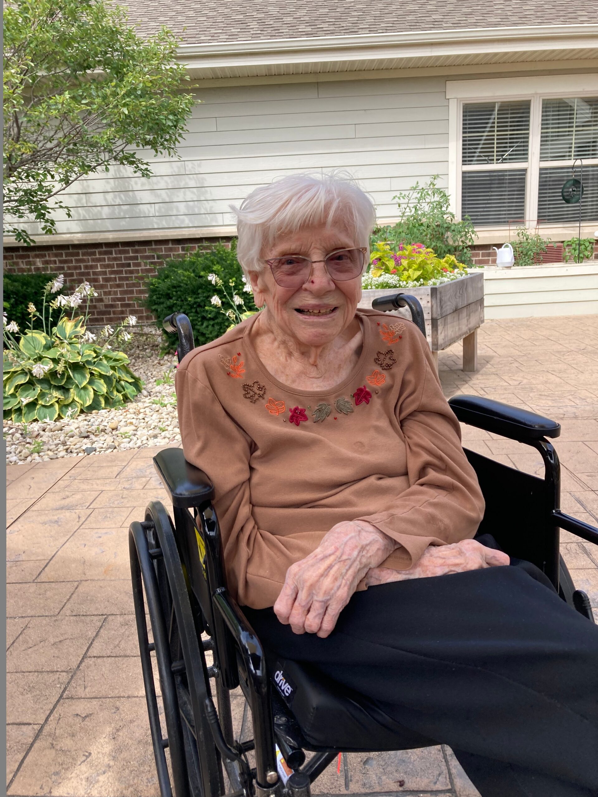 Older woman in a wheelchair outdoors, wearing a brown embroidered top, smiling at the camera with a garden and house in the background.