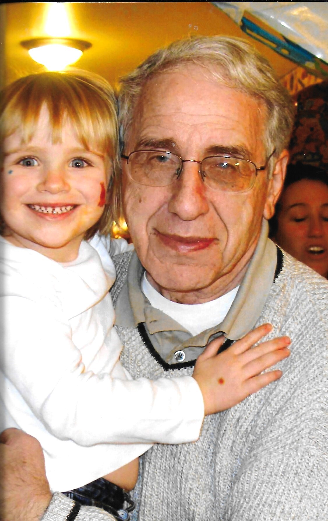 Young girl with face paint smiling and hugging an elderly man wearing glasses at a cozy indoor event.