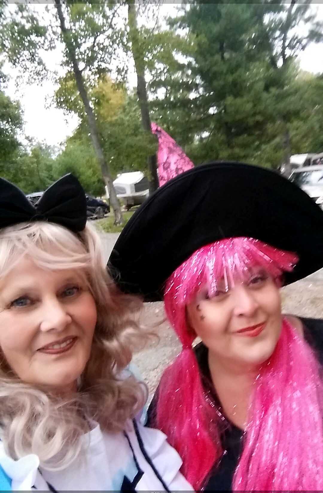 Two women wearing witch hats posing for a selfie outdoors with trees behind them.