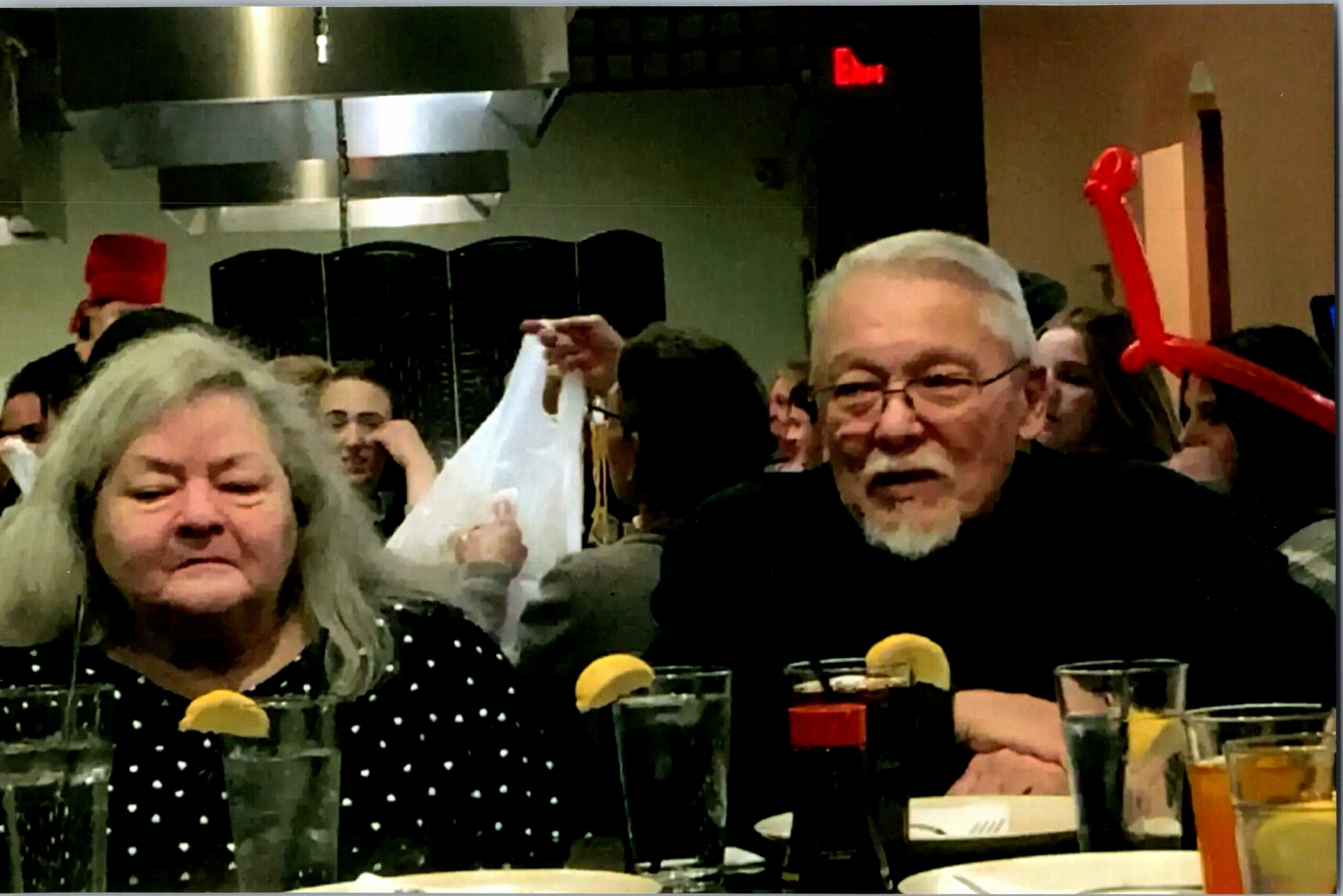 Group of people seated at a restaurant table, drinks with lemon slices, with a man wearing glasses in the foreground and a red balloon in the background.