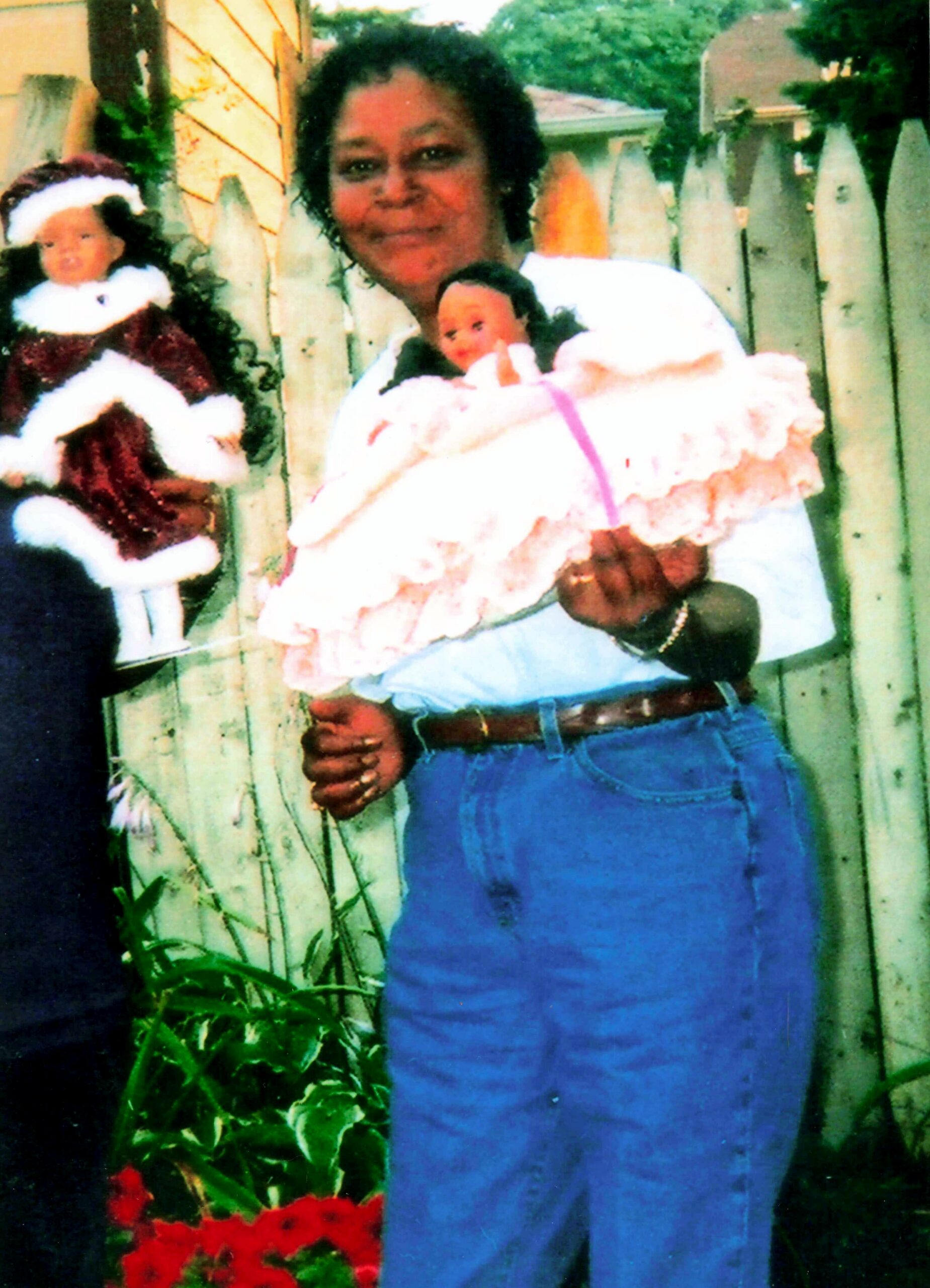 Smiling woman holds a baby wrapped in a pink blanket outdoors beside a wooden fence, with red flowers in the foreground.