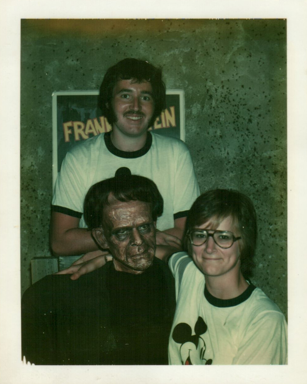 Three friends pose for a retro photo; one person in the front left wears a dark old-man mask while two others smile nearby, with a Frankenstein poster in the background.