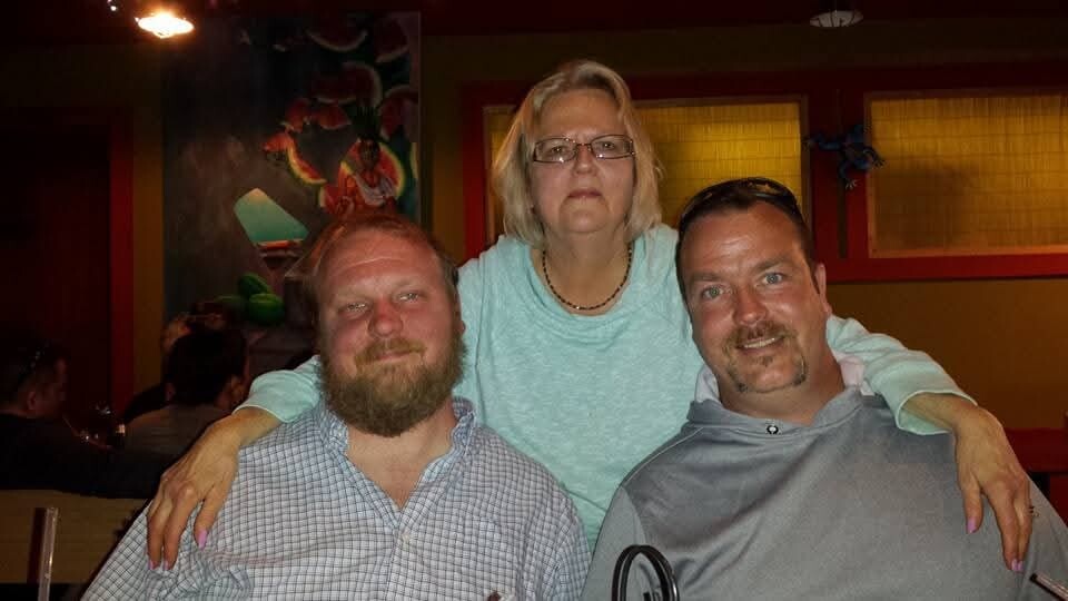 Three adults posing together at a restaurant, with a woman standing behind two men and arms around their shoulders.