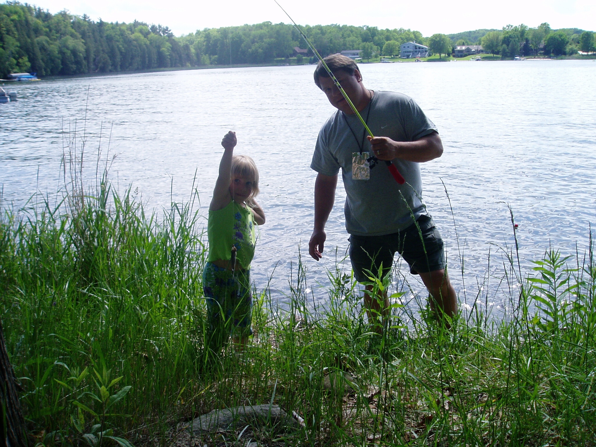Man and child fishing together by a lake, standing among tall grasses with a fishing rod in hand.