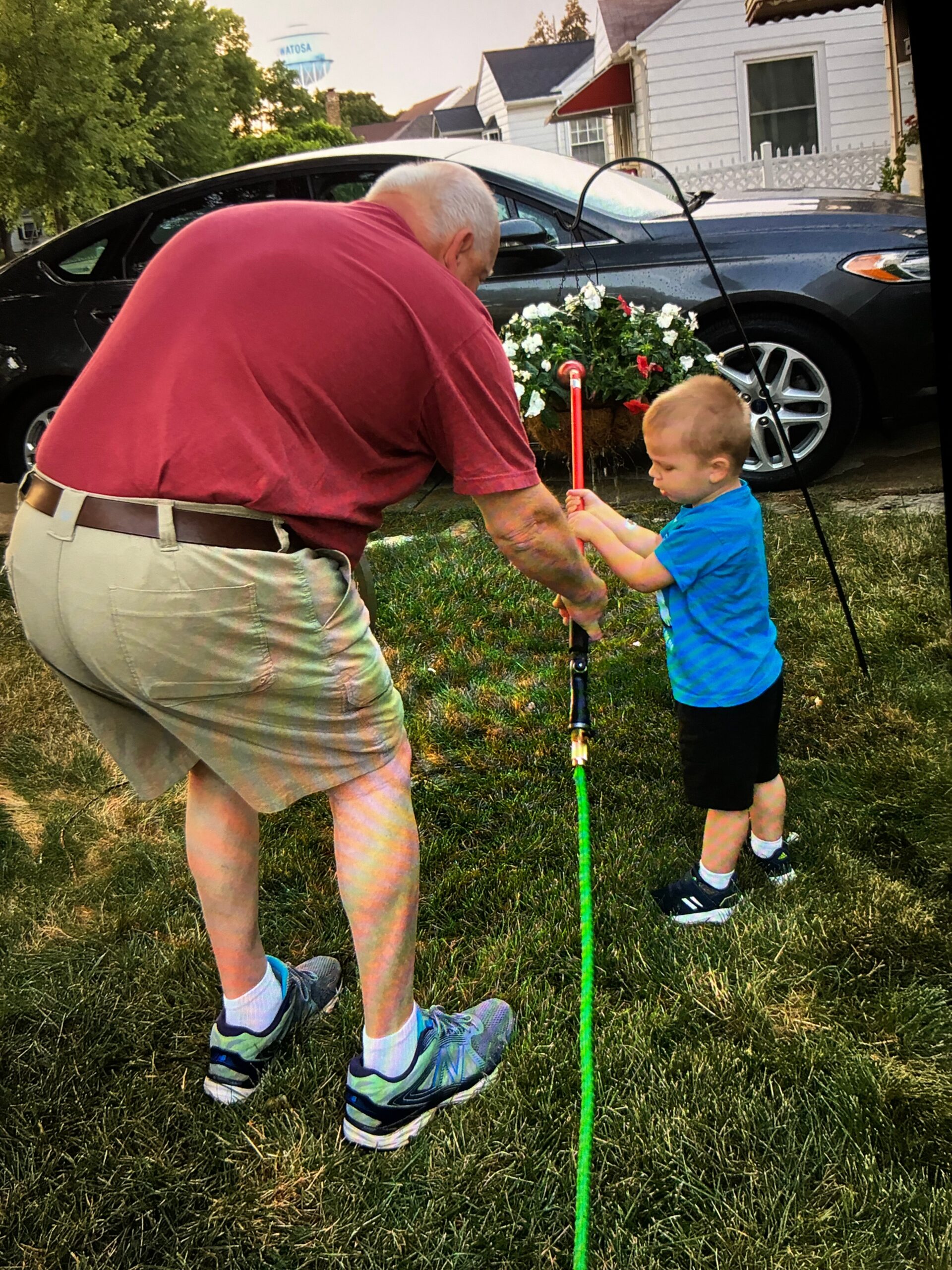 A child in a blue shirt holds a green rope-like pole while an older man in a red shirt assists him outdoors on a grassy yard; a car and houses are in the background.
