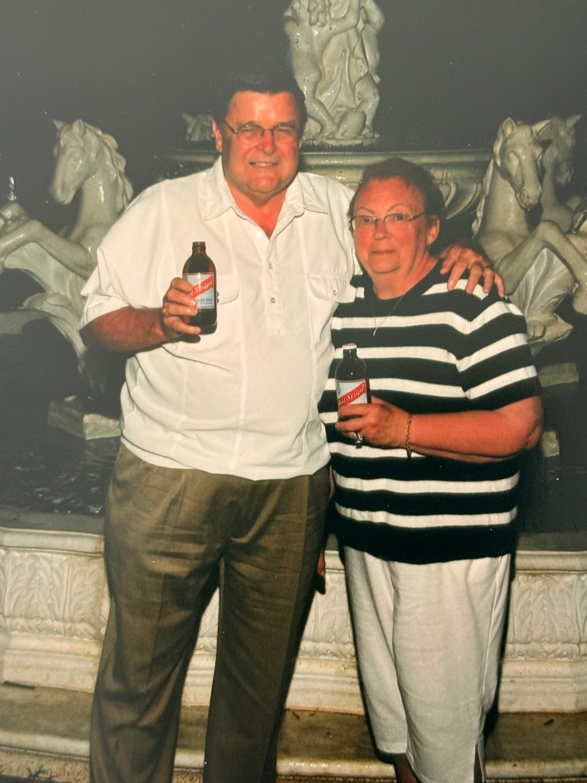 A smiling older couple stands side by side, each holding a bottle of beer, in front of a sculpted horse‑and-figures fountain refreshment pool.