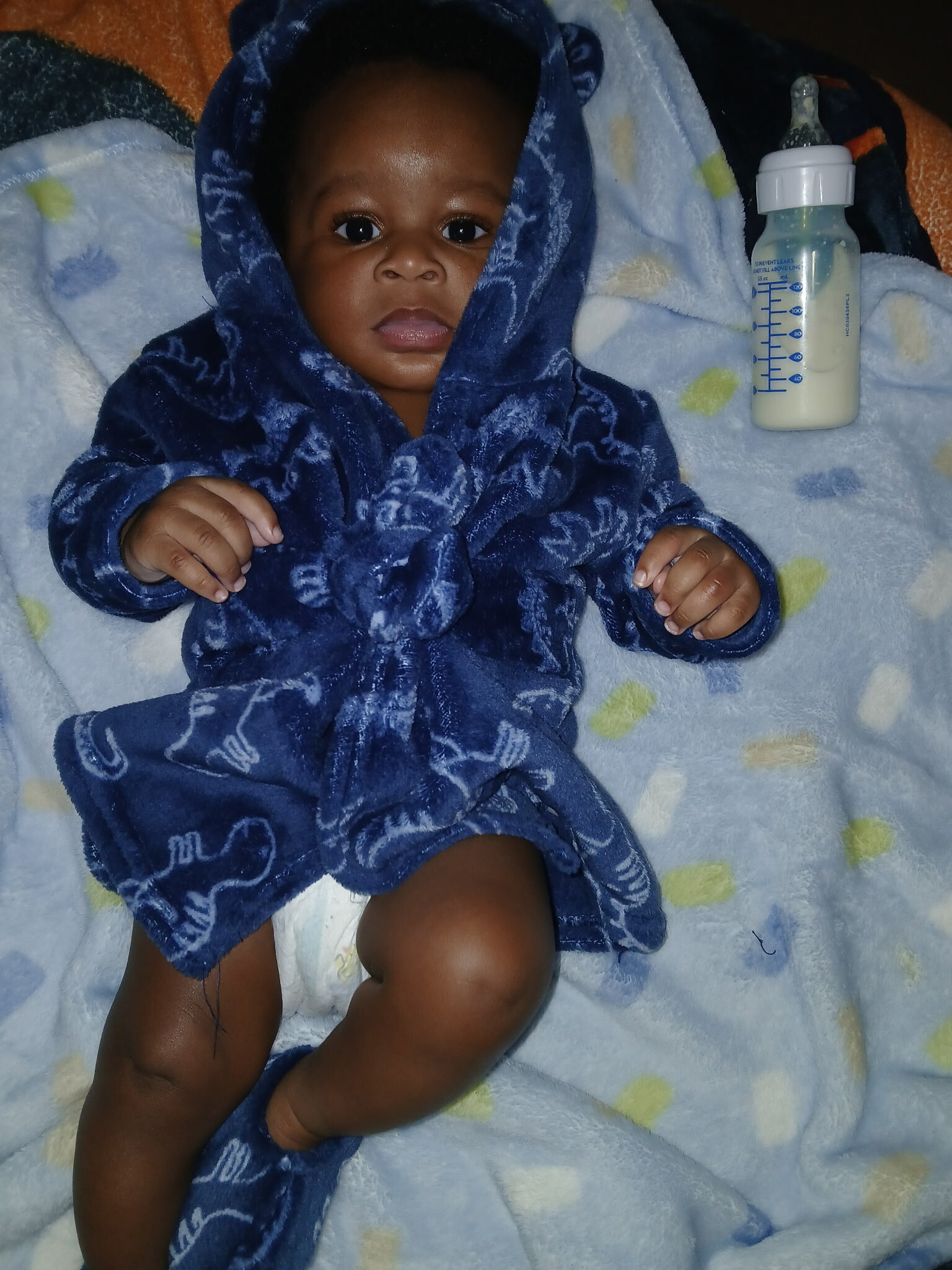 Baby wrapped in a blue towel lies on a pale blanket with a baby bottle nearby.