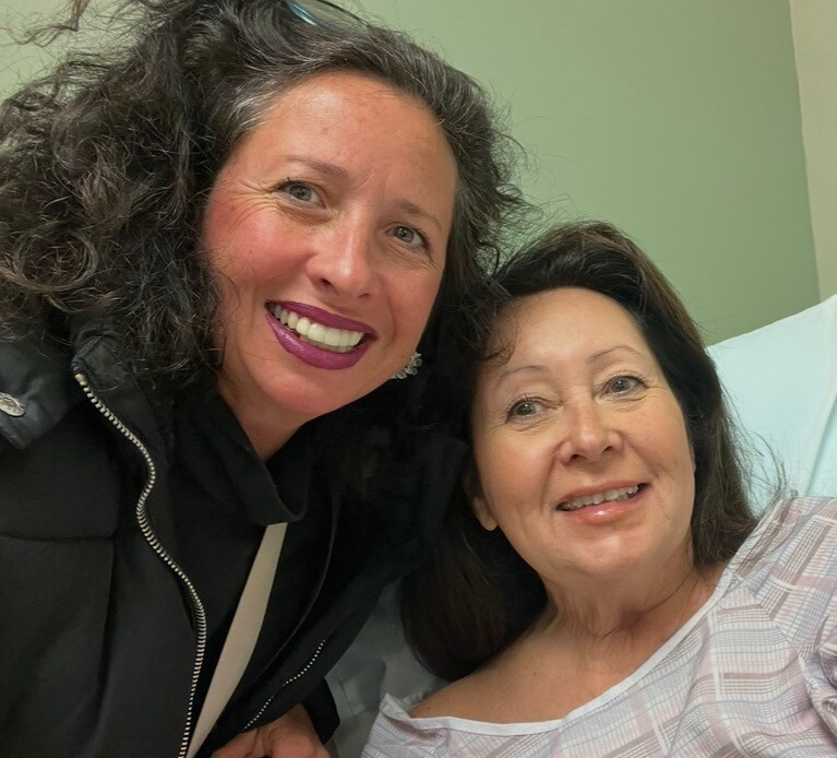 Two women smiling for a selfie in a hospital room; one is wearing a black jacket and the other is a patient in a light hospital gown.