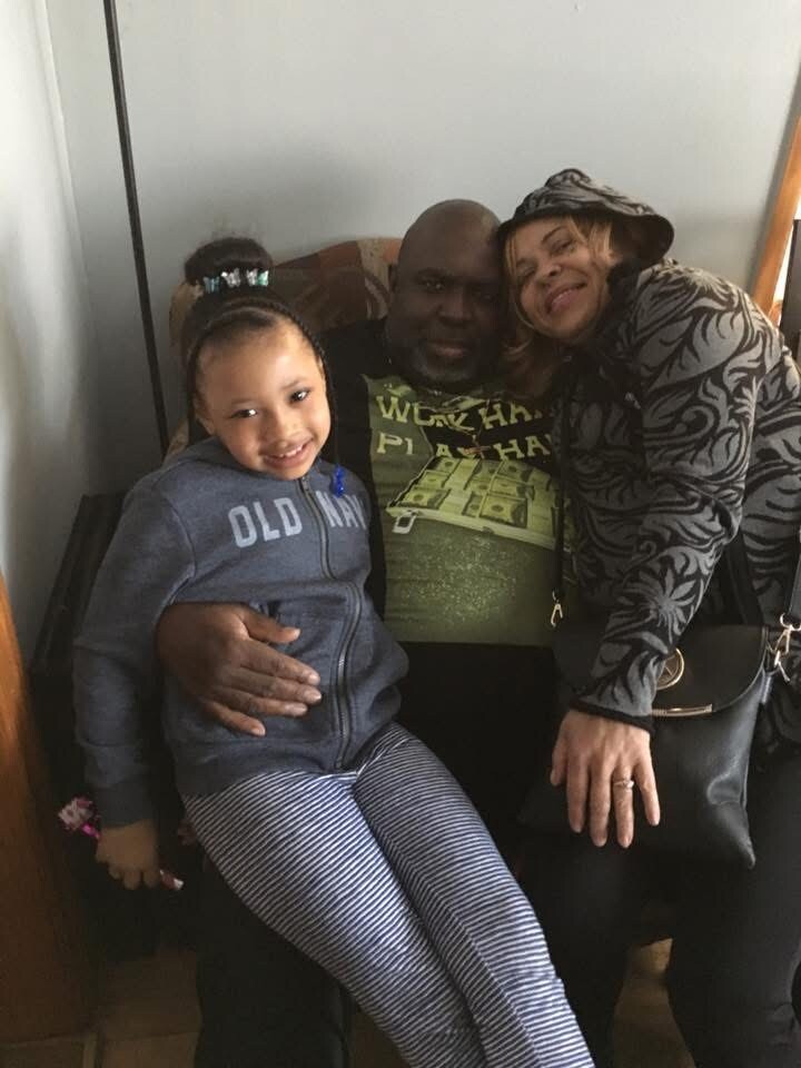 A man sits on a couch with a young girl on his lap, smiling, as a woman in a patterned jacket leans in beside them.
