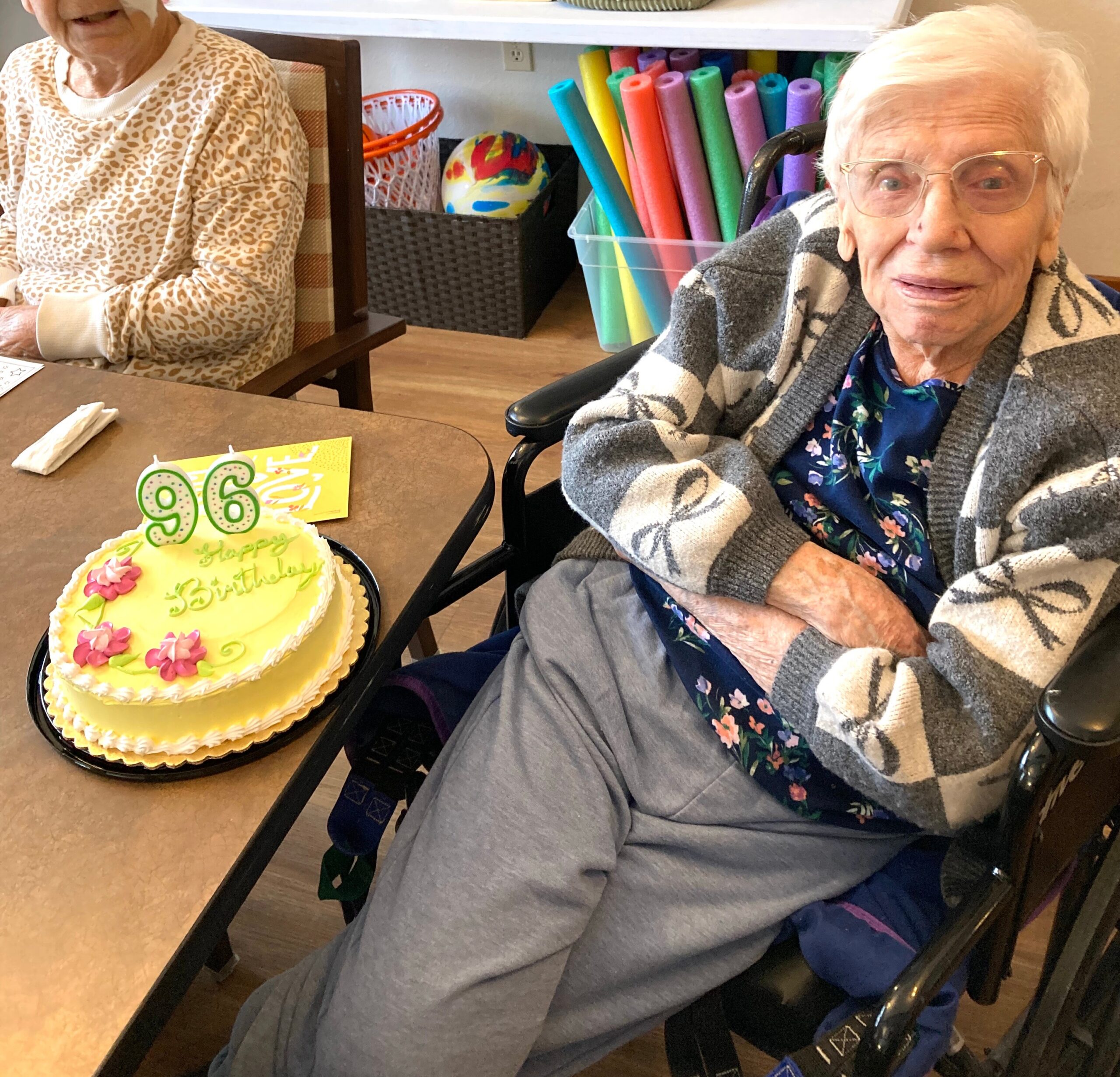 Elderly person in a wheelchair smiling next to a birthday cake with large green '96' candles on a table by another seated person in a leopard-patterned sweater.
