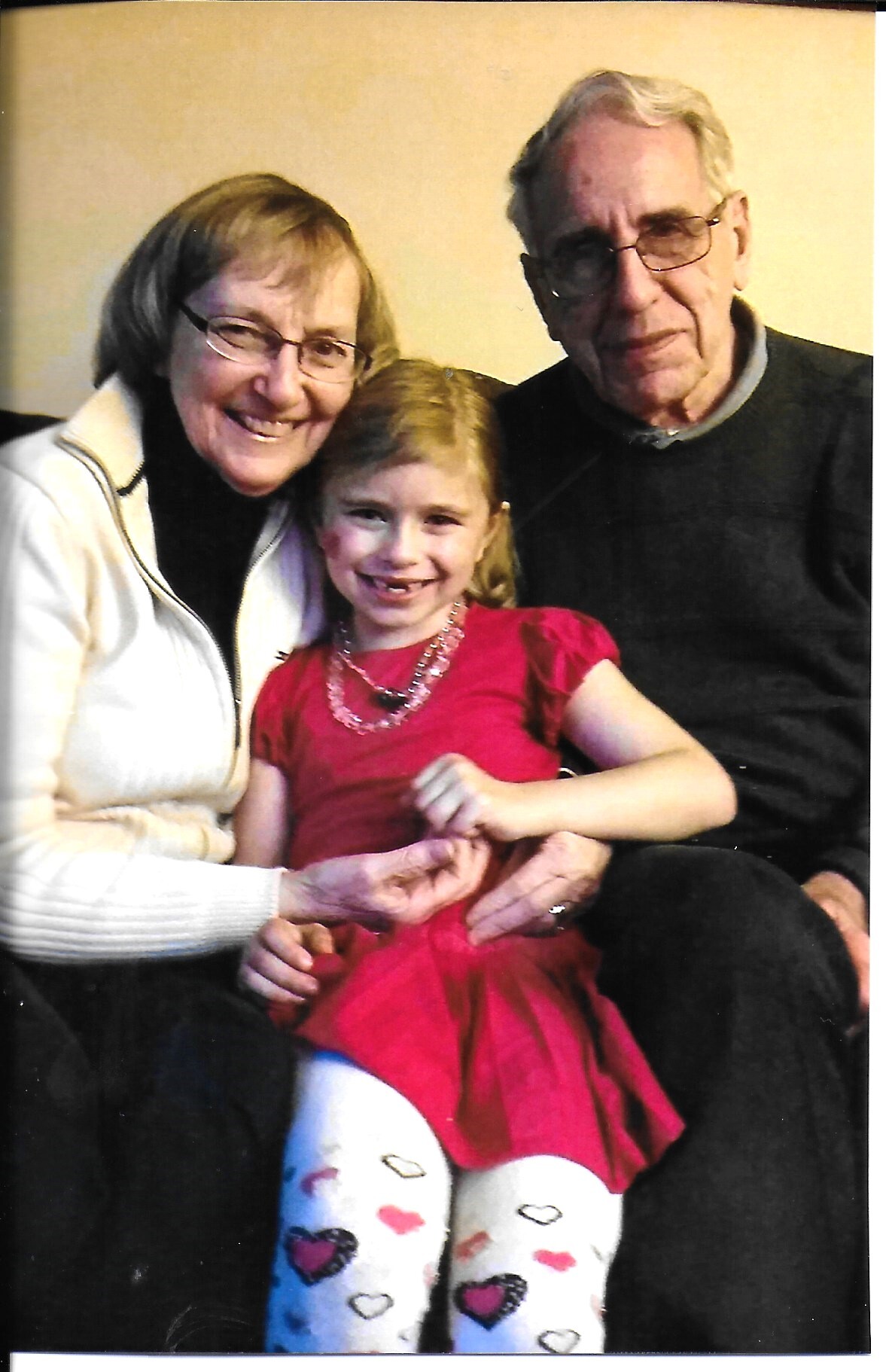 Grandmother and grandfather sit with their smiling granddaughter in a red dress on a couch.