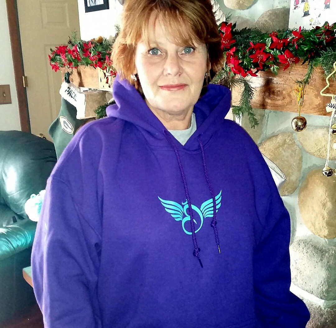 Portrait of a woman with short red hair wearing a purple hoodie with a blue winged emblem, indoors with holiday garlands in the background.