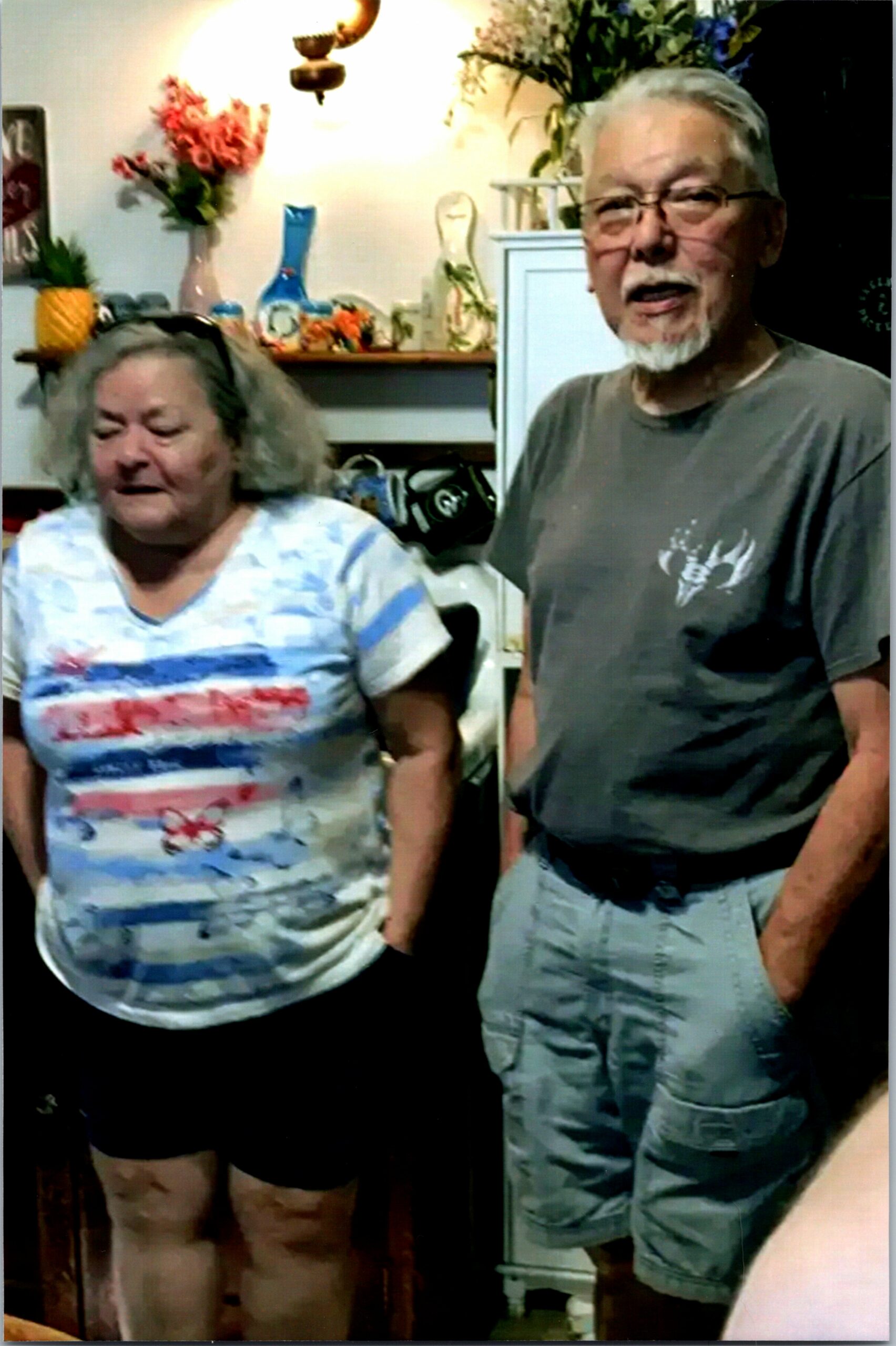 Two older adults standing side by side indoors: a woman in a patterned shirt on the left and a man in a gray shirt and cargo shorts on the right, with shelves and flowers in the background.