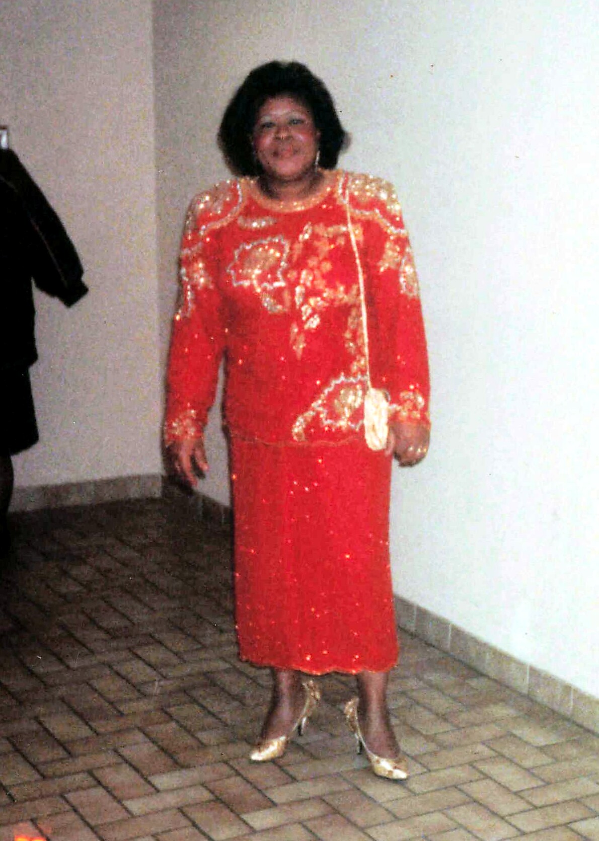 Smiling woman in a bright red, sequined traditional dress with gold embroidery, standing indoors on a brick floor with heels and a small purse at her side.