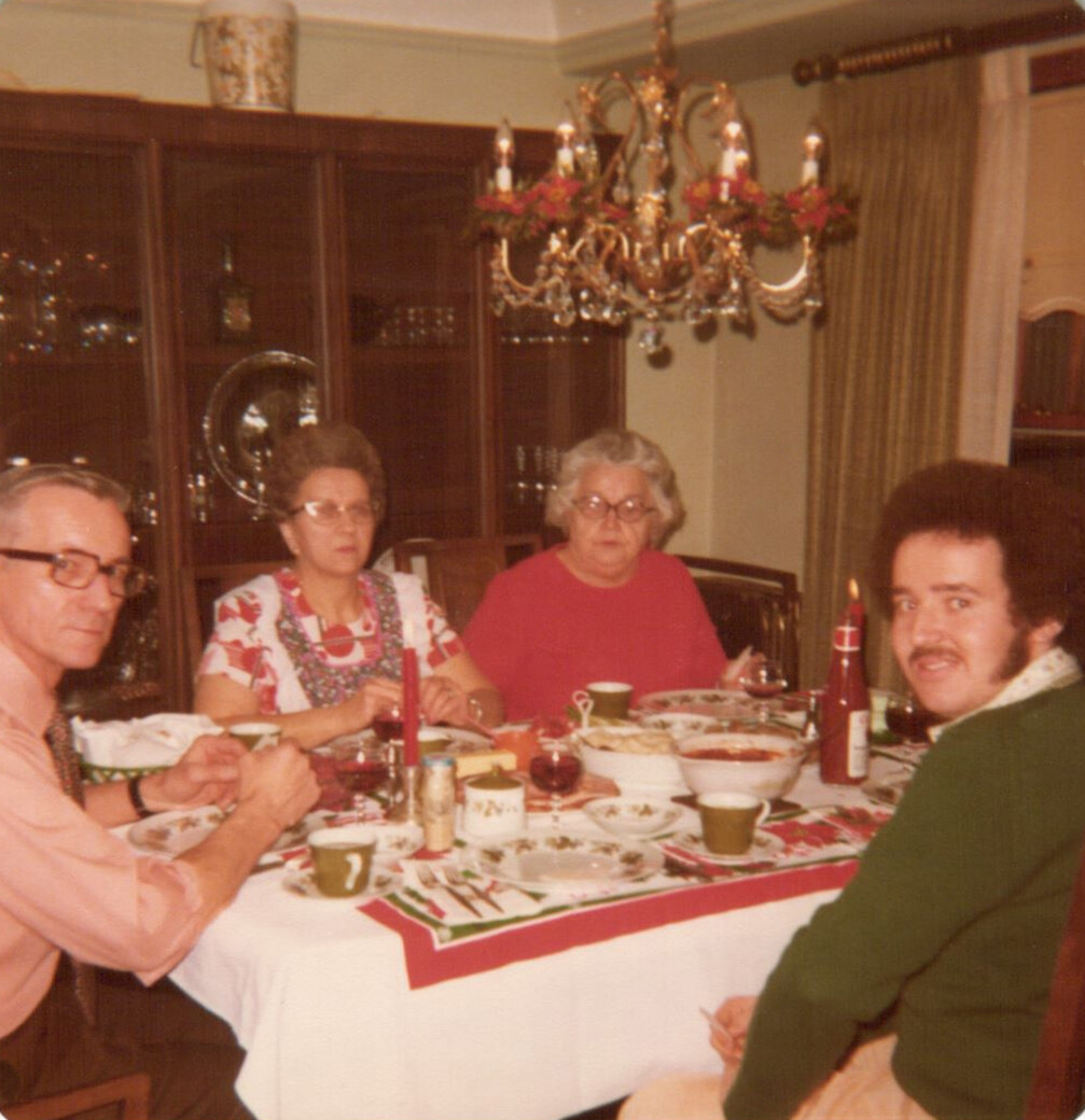 Four people sit around a dining table for a holiday meal, with a chandelier overhead and festive tableware.