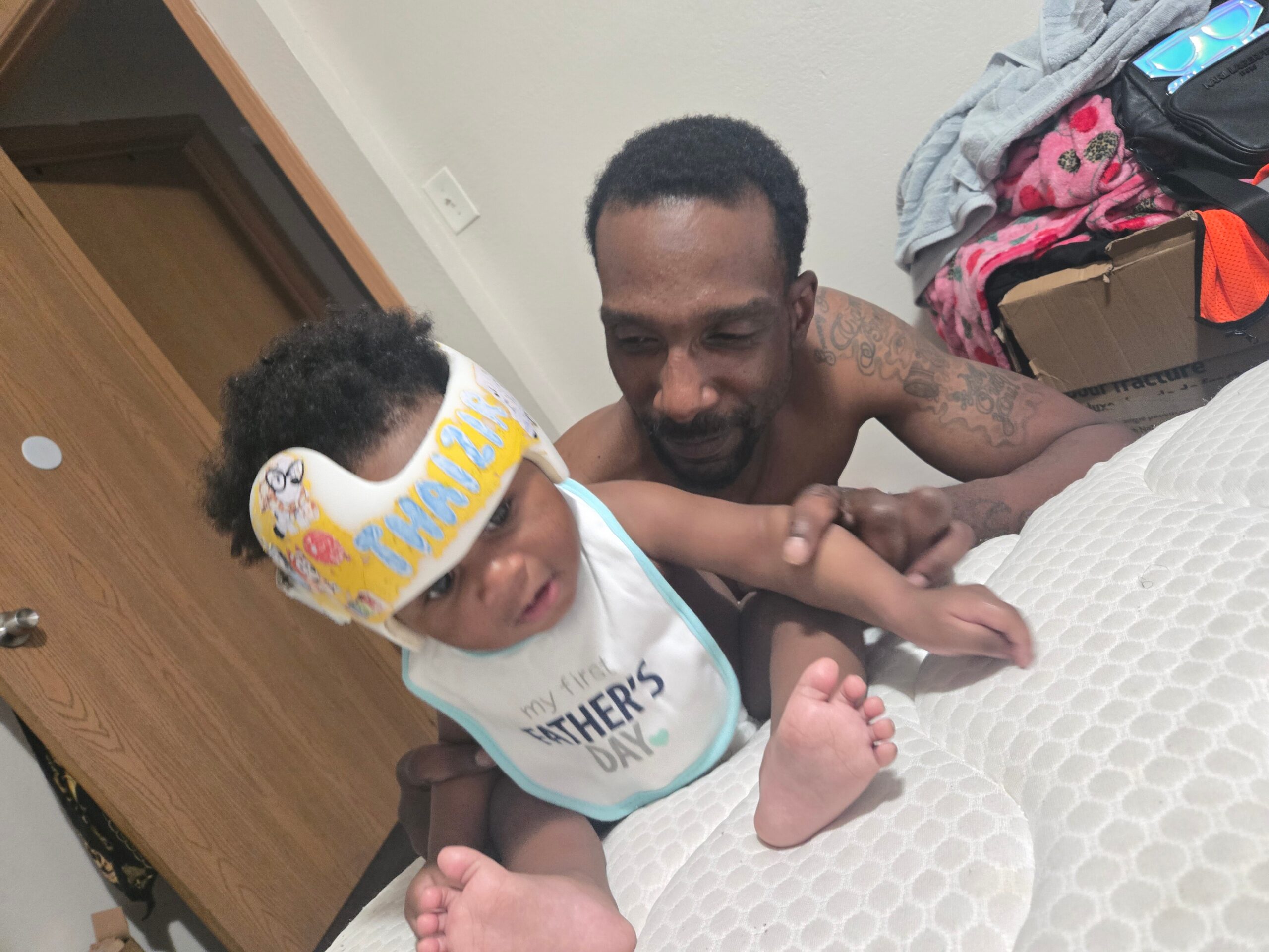 Shirtless man and baby lying on a bed, baby wearing a yellow helmet and bib that says 'My First Father's Day'.