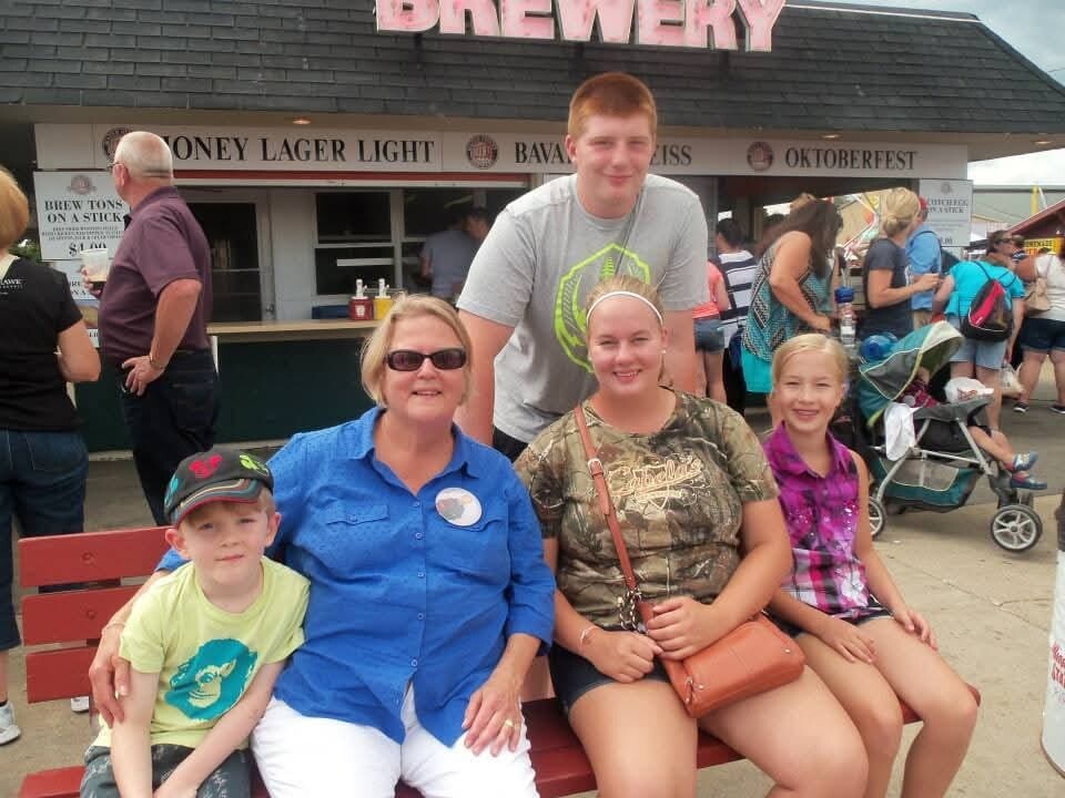 Family posing on a red bench in front of a brewery stand, with a boy wearing a cap and a woman in a blue shirt smiling at the camera.