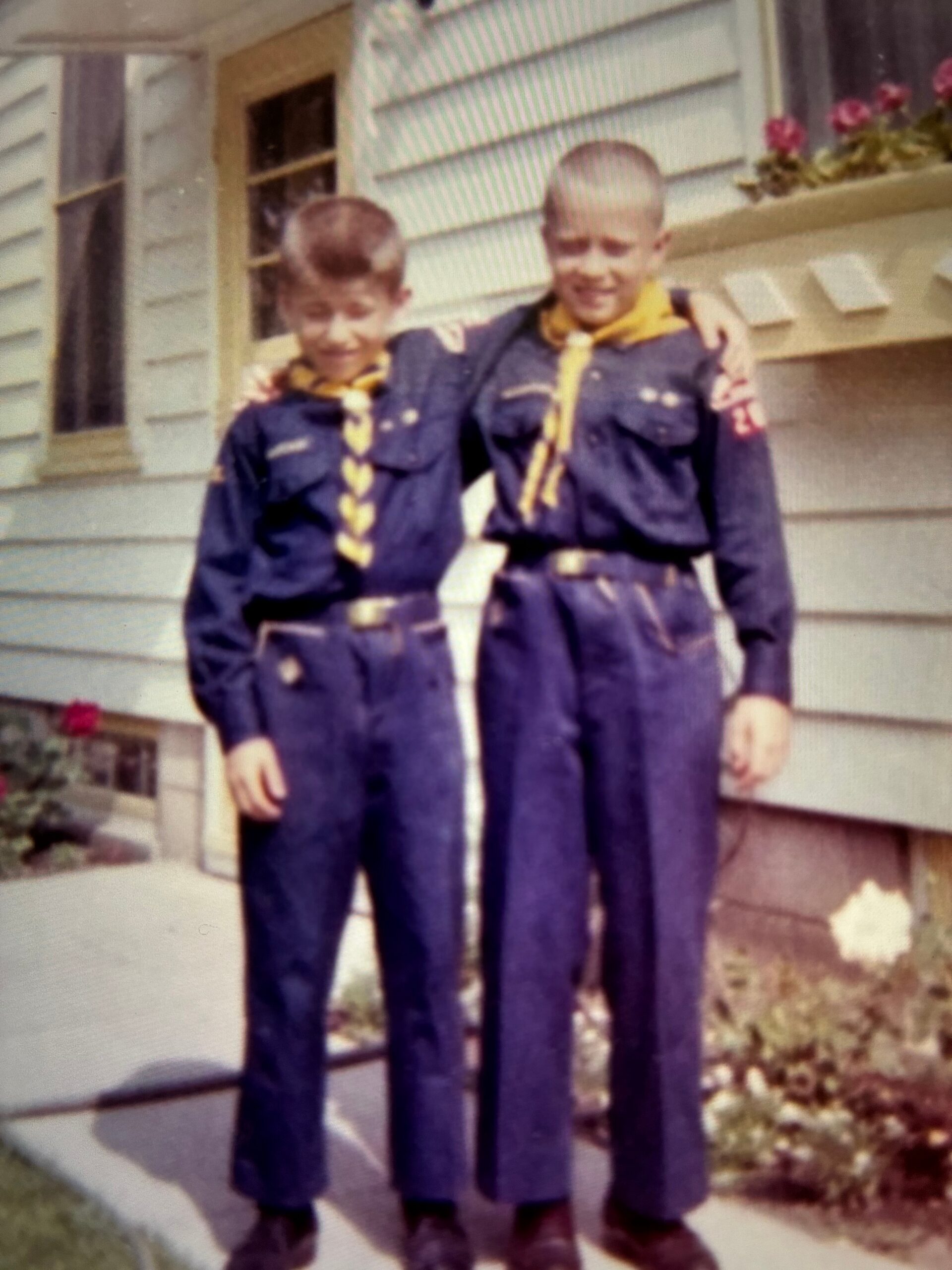 Two boys in navy Cub Scout uniforms with yellow neckerchiefs pose with arms around each other in front of a house.