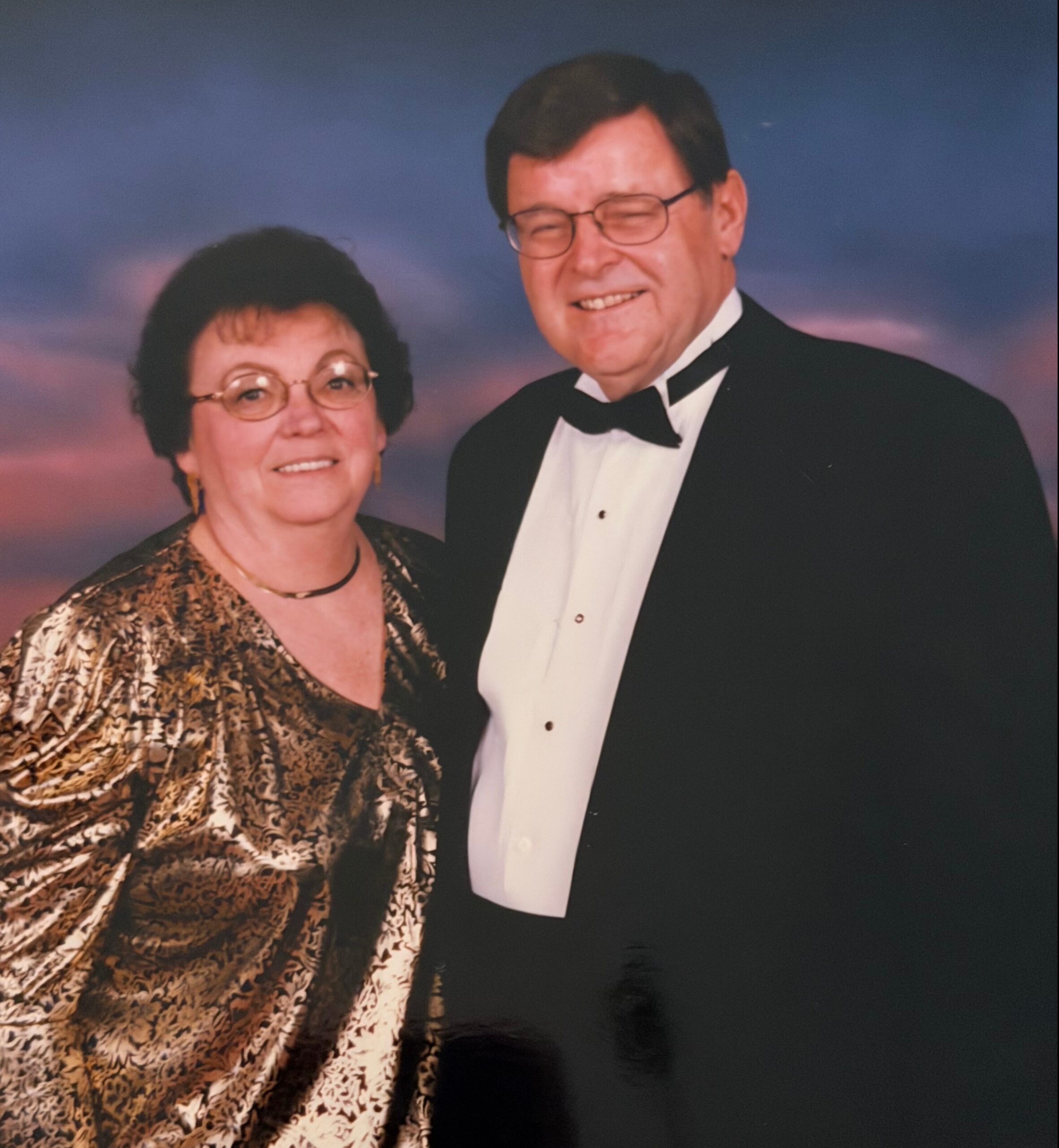 Smiling elderly couple posing together in formal attire against a sunset backdrop