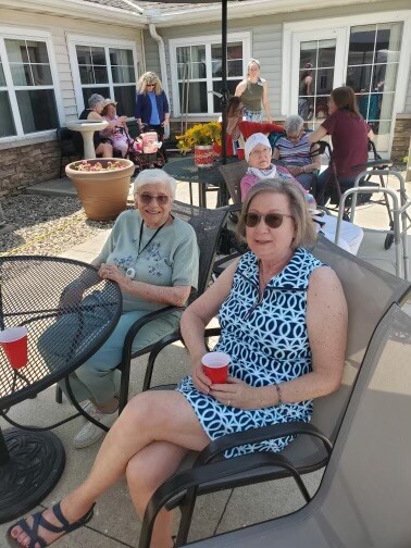 Older adults sit around outdoor tables on a sunny patio, chatting and holding red cups during a social gathering at a residential facility.
