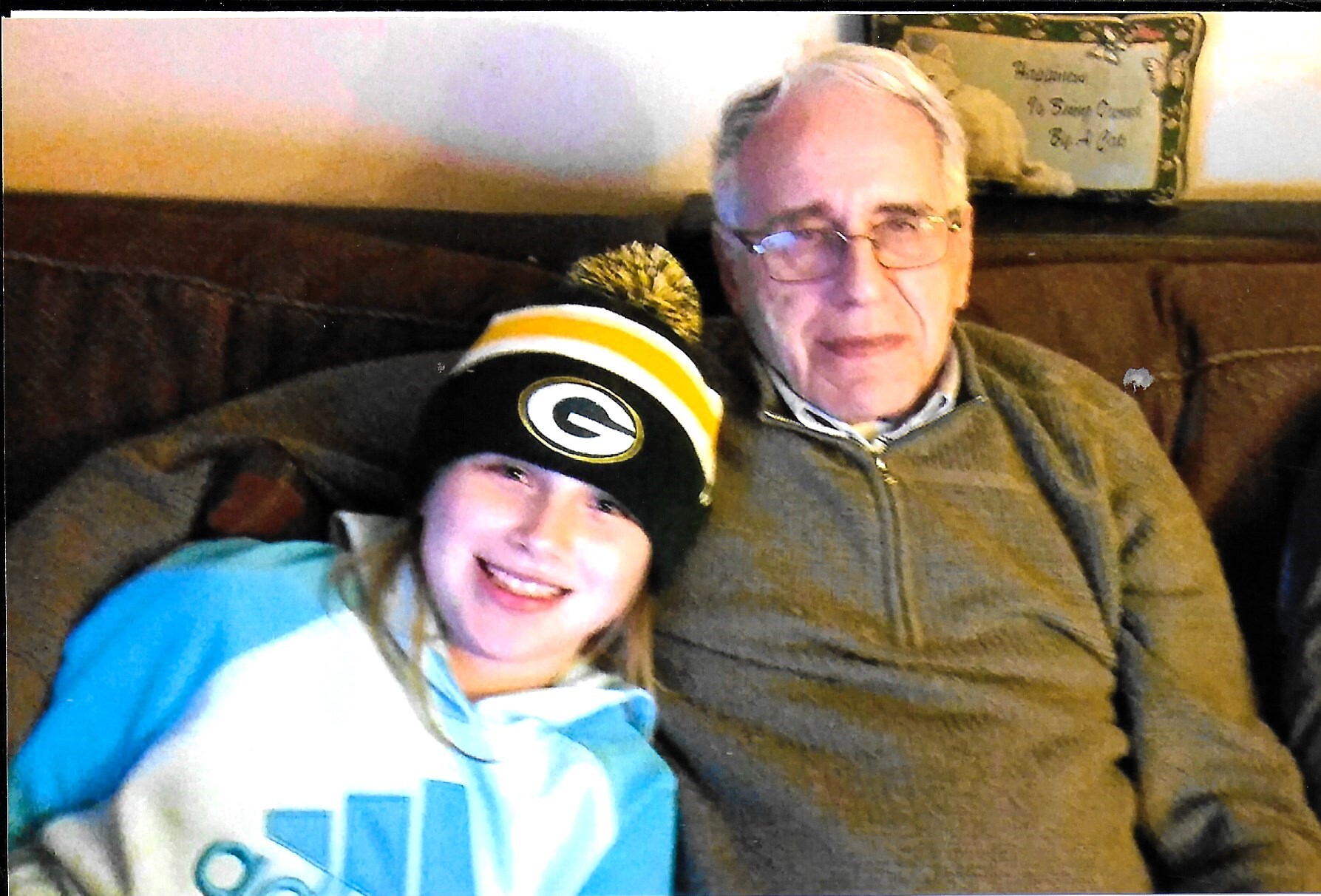 Smiling girl in a black and yellow Packers beanie with a pom-pom, leaning against an older man wearing a brown zip-up sweater on a couch.