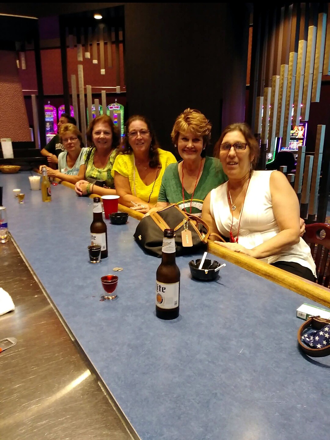 Five women sit along a blue bar in a casino, smiling at the camera with drinks and a purse on the counter and slot machines in the background.