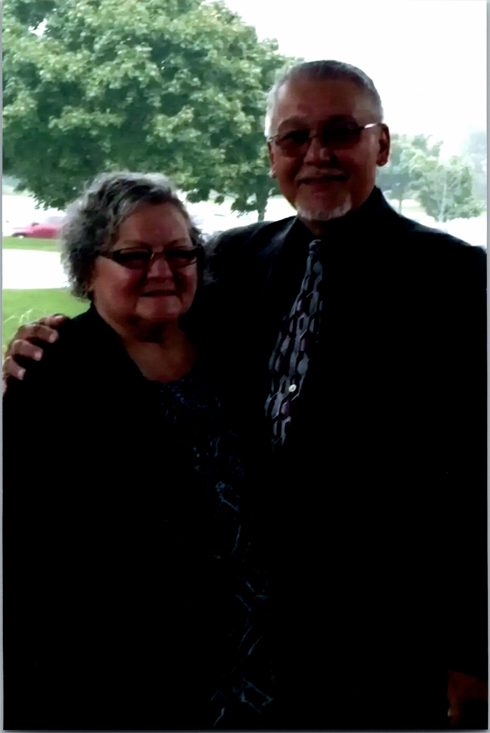 Two adults posing together outdoors with trees in the background; man in a suit and patterned tie, woman in a dark toped outfit.