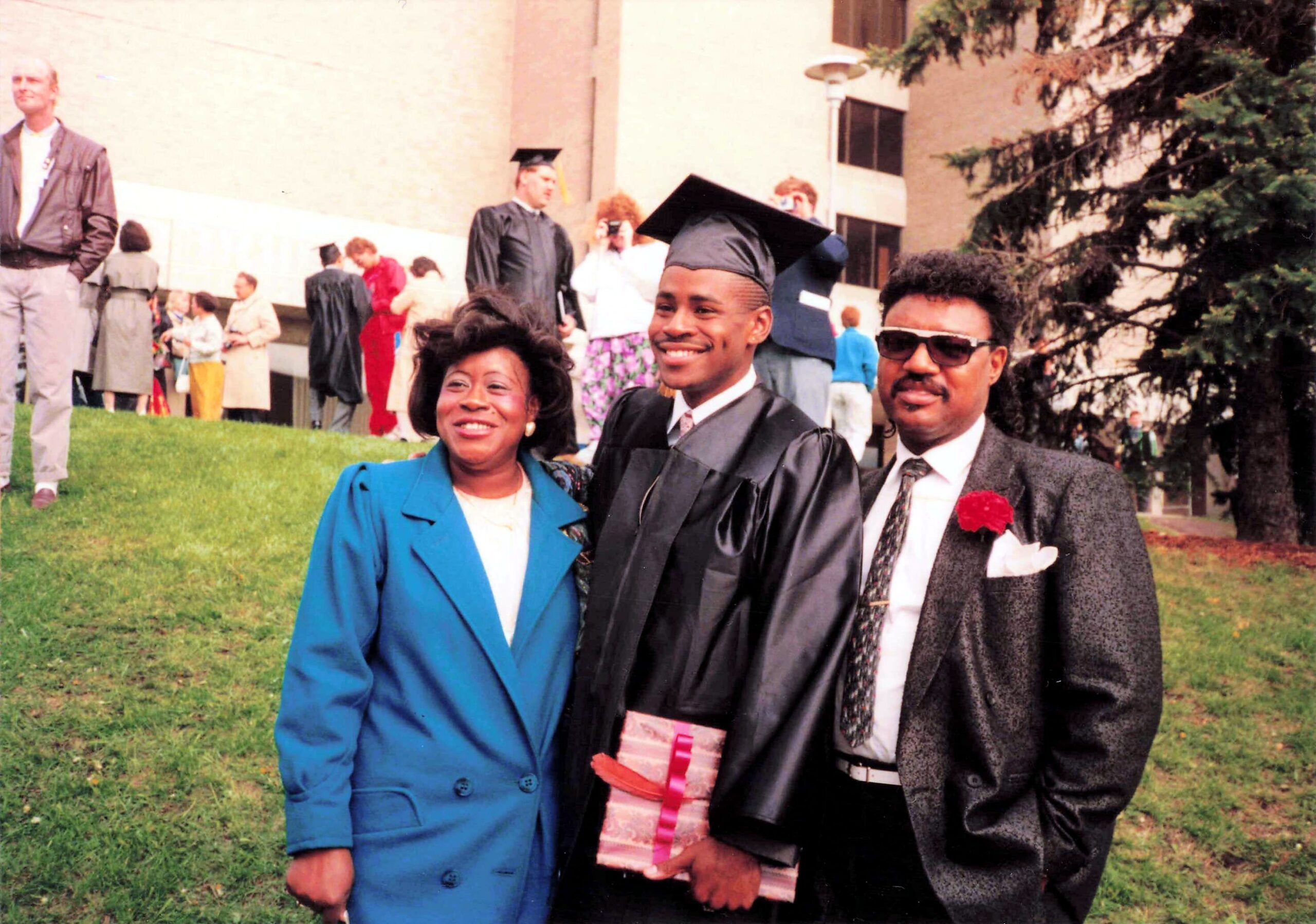 Three people pose at a graduation ceremony outdoors: a smiling woman in a bright blue suit, a graduate in cap and gown, and a man in a suit with sunglasses holding a diploma sleeve, with classmates and a campus building in the background.