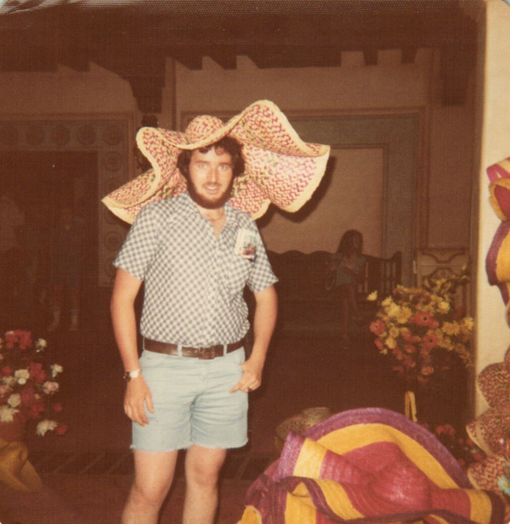 Man in a checkered shirt and light shorts poses indoors, wearing an oversized sombrero for a playful look on camera.