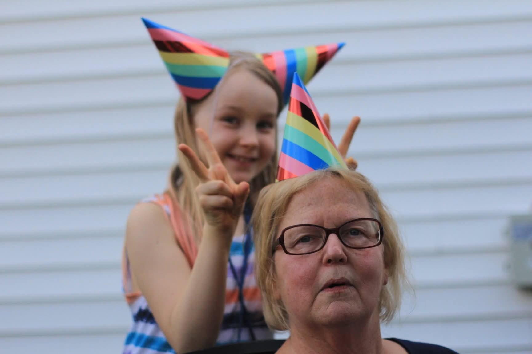 Girl smiling and making a peace sign from behind an older woman wearing rainbow party hats at a celebration, with a light-colored wall in the background.