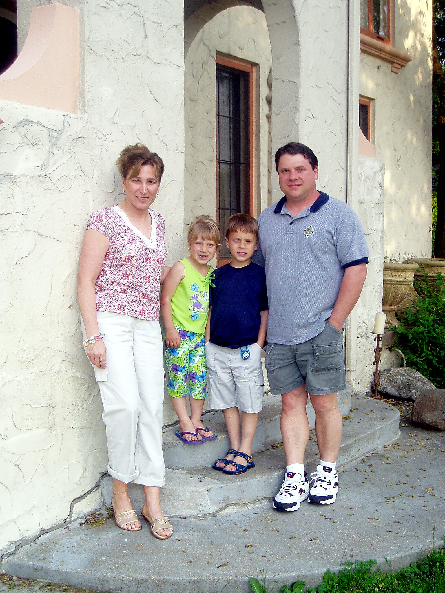 Family of four posing together on the stone steps outside a light-colored stucco house with an arched doorway behind them