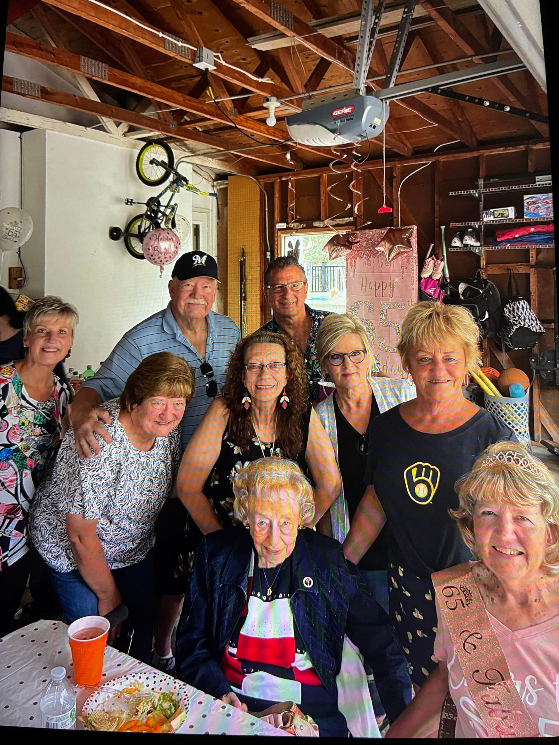 Group of smiling adults at a birthday party in a garage, with balloons, a banner, and snacks on a table.