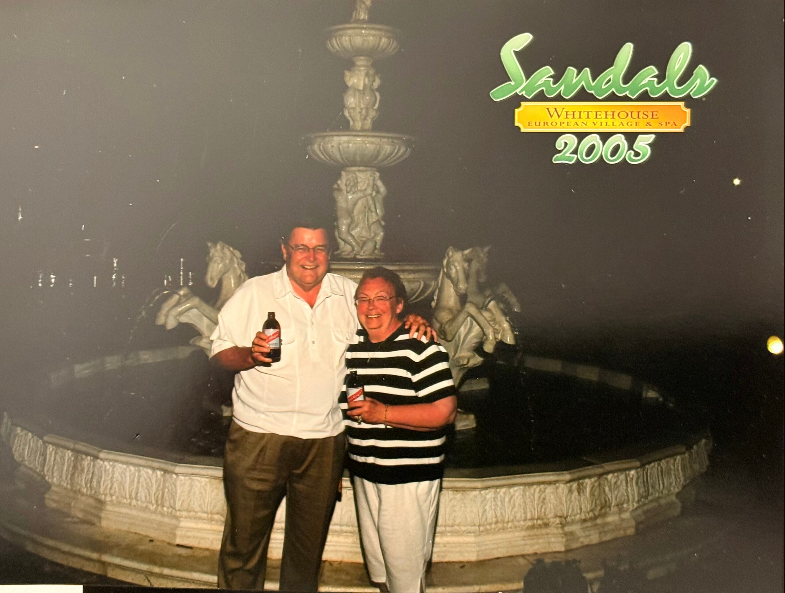 Smiling couple posing at night in front of a large ornate fountain with a Sandals 2005 sign, each holding a beer bottle.