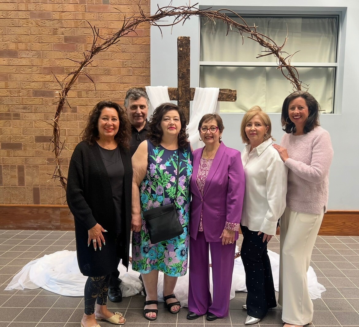 Six adults posing for a photo indoors under a rustic wooden arch with a cross and white fabric backdrop, everyone smiling.