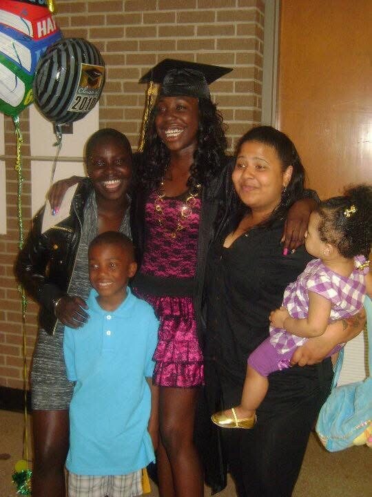 Group of five people posing for a graduation photo, with a graduate in cap and gown at center, flanked by family members and a child, balloons in the background.