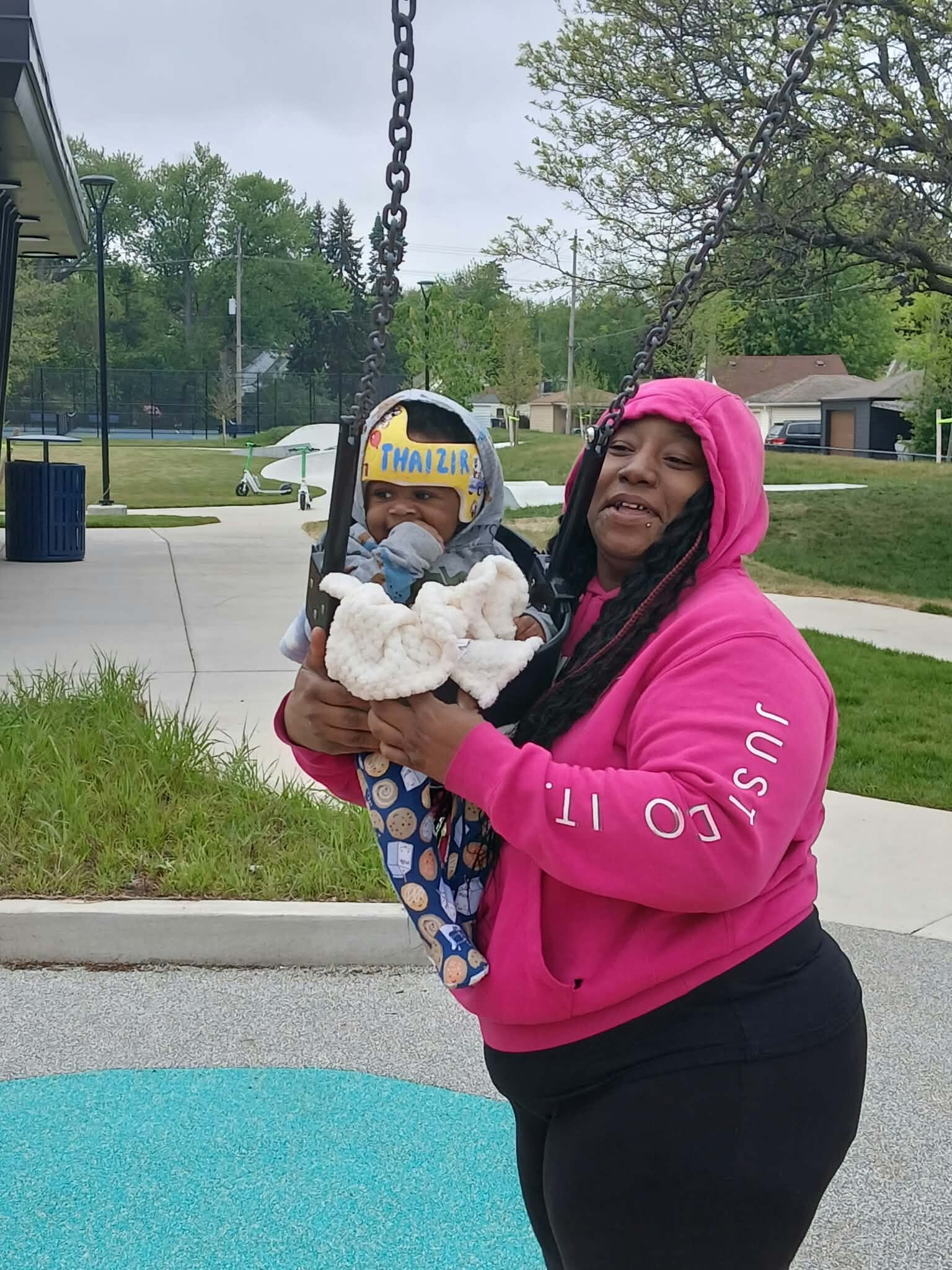 Woman in a pink hoodie pushing a baby on a swing at a park, the baby wearing a light helmet with colorful writing on it.