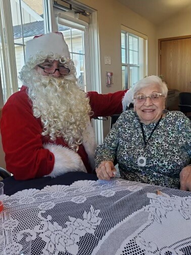 Santa Claus with white beard sits beside an elderly woman at a lace-covered table, smiling indoors.