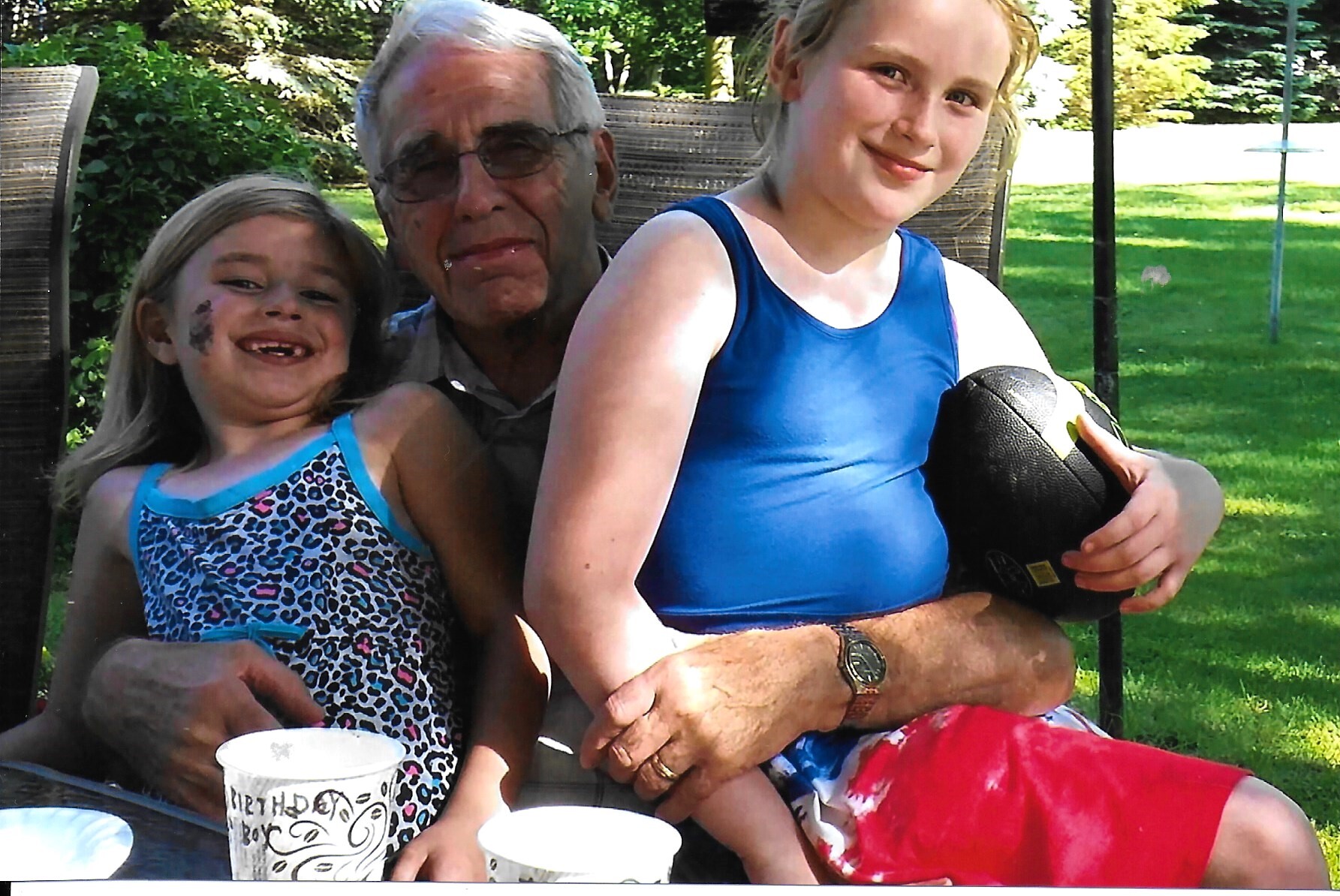 Three generations posing outdoors: an elderly man sits between two smiling girls, one with missing teeth on the left and a girl in blue holding a basketball on the right.