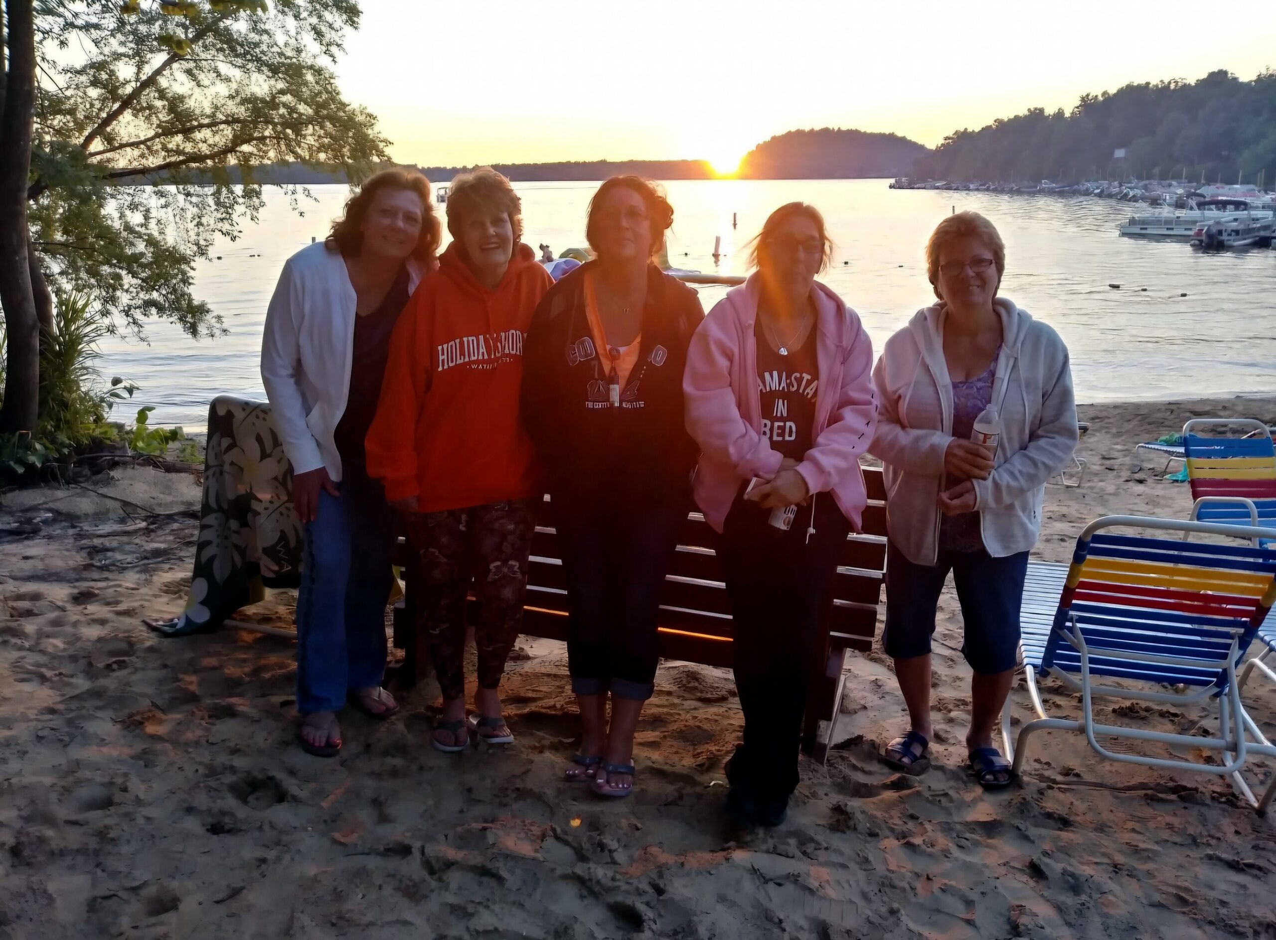 Five women posing on a sandy beach at sunset with a calm lake and boats in the background.