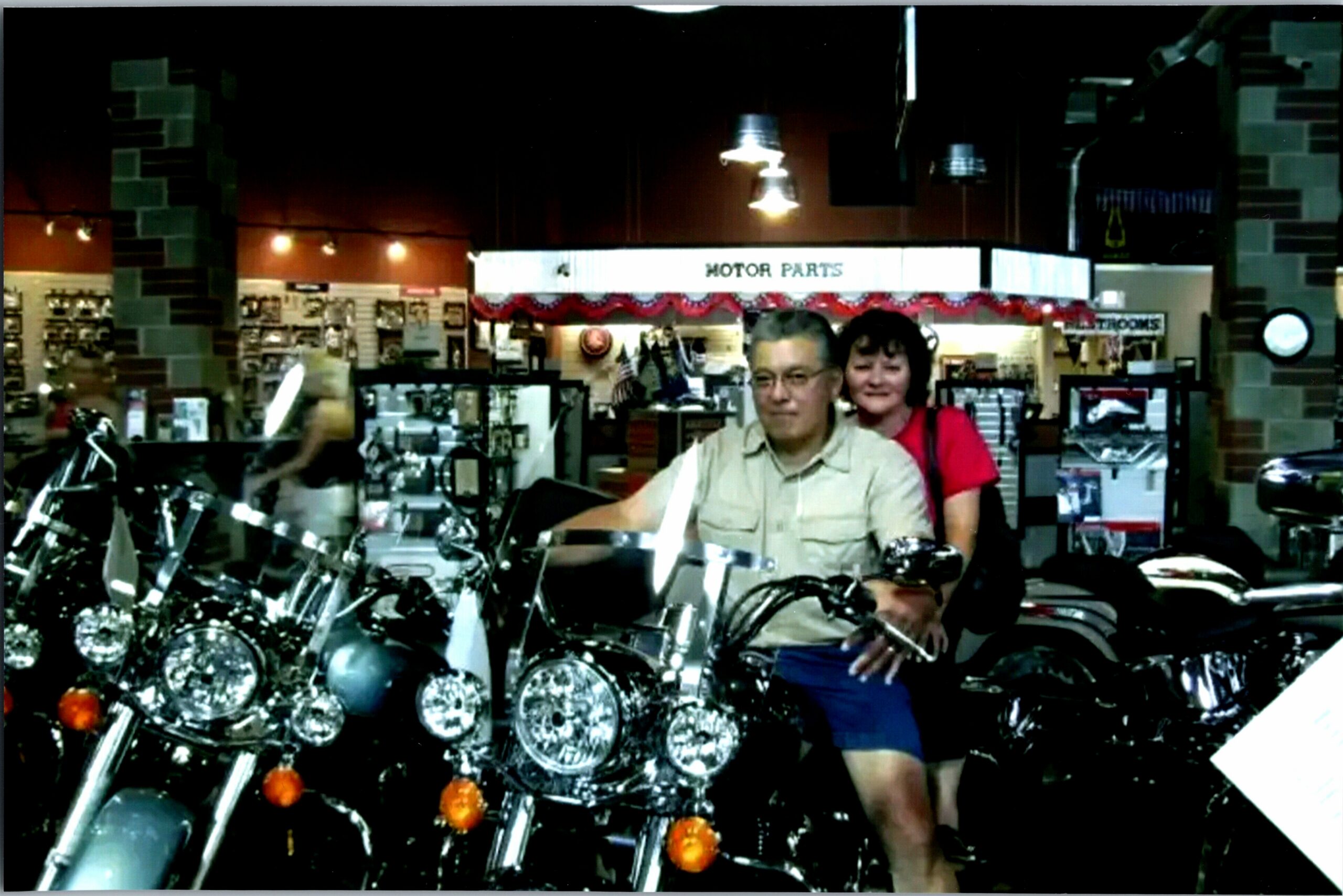 Smiling couple posing in a motorcycle showroom with chrome bikes and a 'Motor Parts' sign in the background at night.