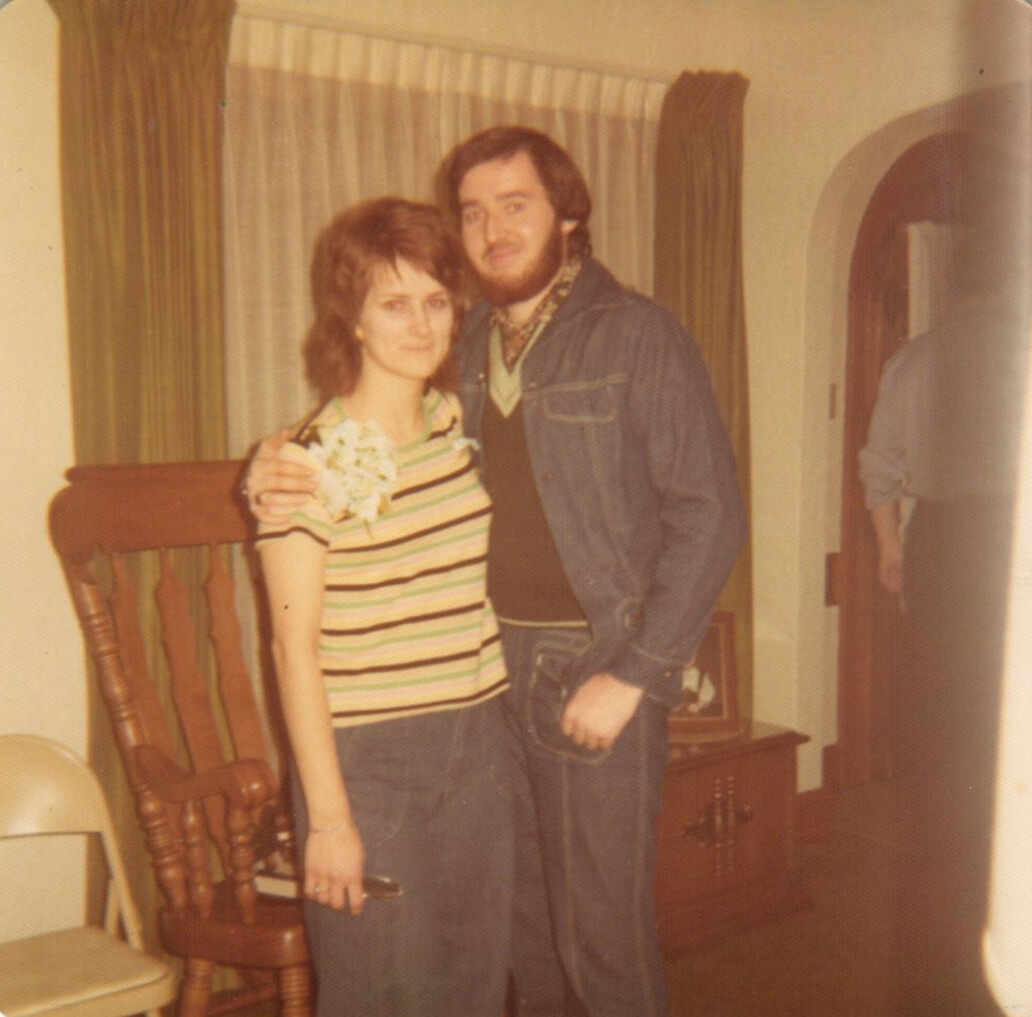 Young couple posing together in a living room; woman wears a striped shirt with a corsage, man in a denim jacket and sweater.